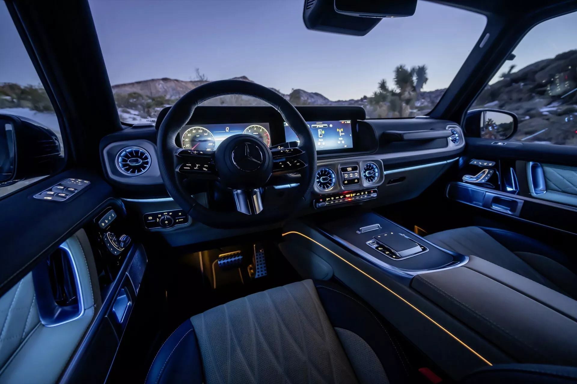 Interiérový pohľad na Mercedes-Benz G-Klasse 2024, zdôrazňujúci dizajn kokpitu z pohľadu zadného pasažiera, predstavujúci multifunkčný športový volant, palubnú dosku s digitálnym prístrojovým panelom, širokouhlý informačný systém, ovládanie klímy, ambientné interiérové osvetlenie, luxusné predné sedadlá, volič prevodovky, držiaky nápojov, dizajn dverí s elektrickými ovládaniami a prémiové materiály dokončenia.