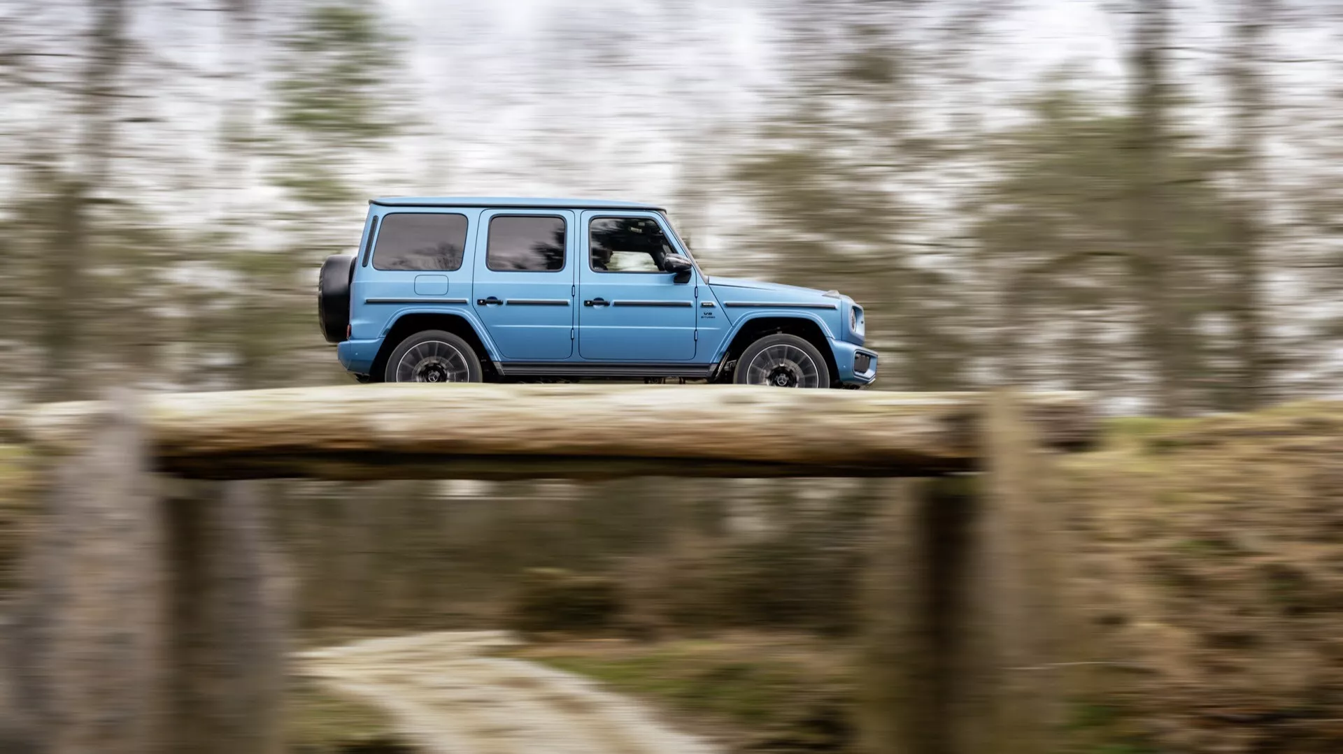 Pohybujúce sa Mercedes-Benz G-Klasse (Trieda G) 465 z roku 2024 na nezpevnenej ceste, zachytené technikou panning pre zdôraznenie pohybu, zobrazuje profilový pohľad zdôrazňujúci off-road dizajn a zliatinové kolesá.