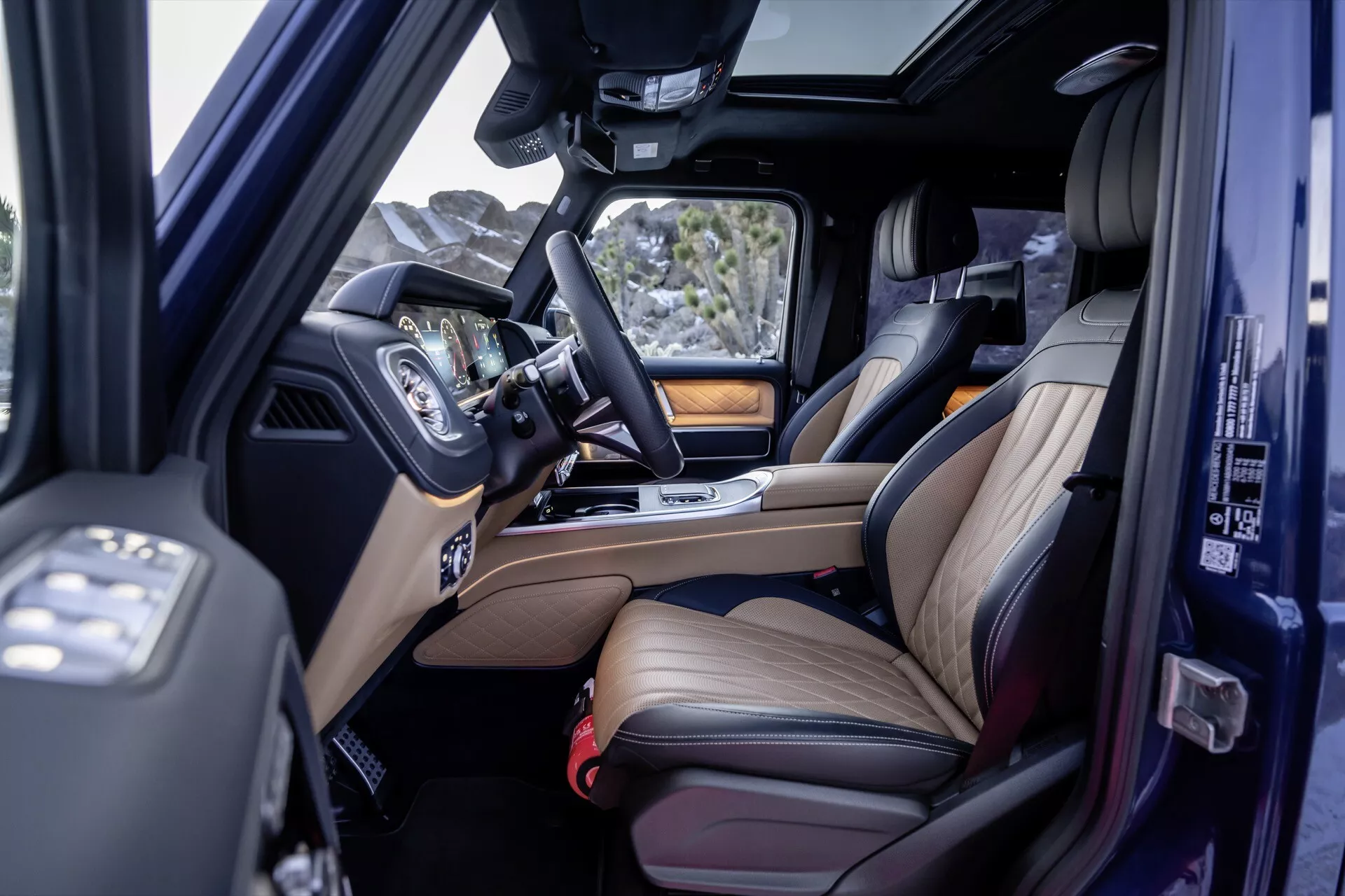 Interiérový pohľad na Mercedes-Benz G-Class 2024 s prednými a zadnými koženými sedadlami s drevitými akcentmi, infotainmentom a strešným oknom z zvýšeného uhla