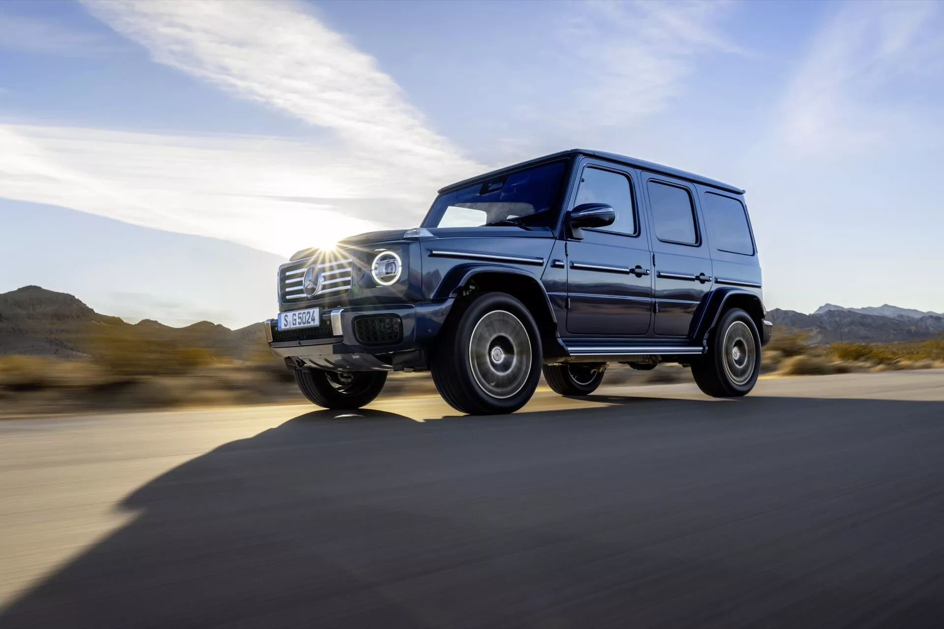 Luxusné SUV Mercedes-Benz G-Class 465 ročník 2024 v dynamickom bočnom pohľade idúce po asfaltovej ceste, s LED prednými svetlami, robustným dizajnom kolies, vysokou svetlou výškou a lesklým povrchom na dennom svetle