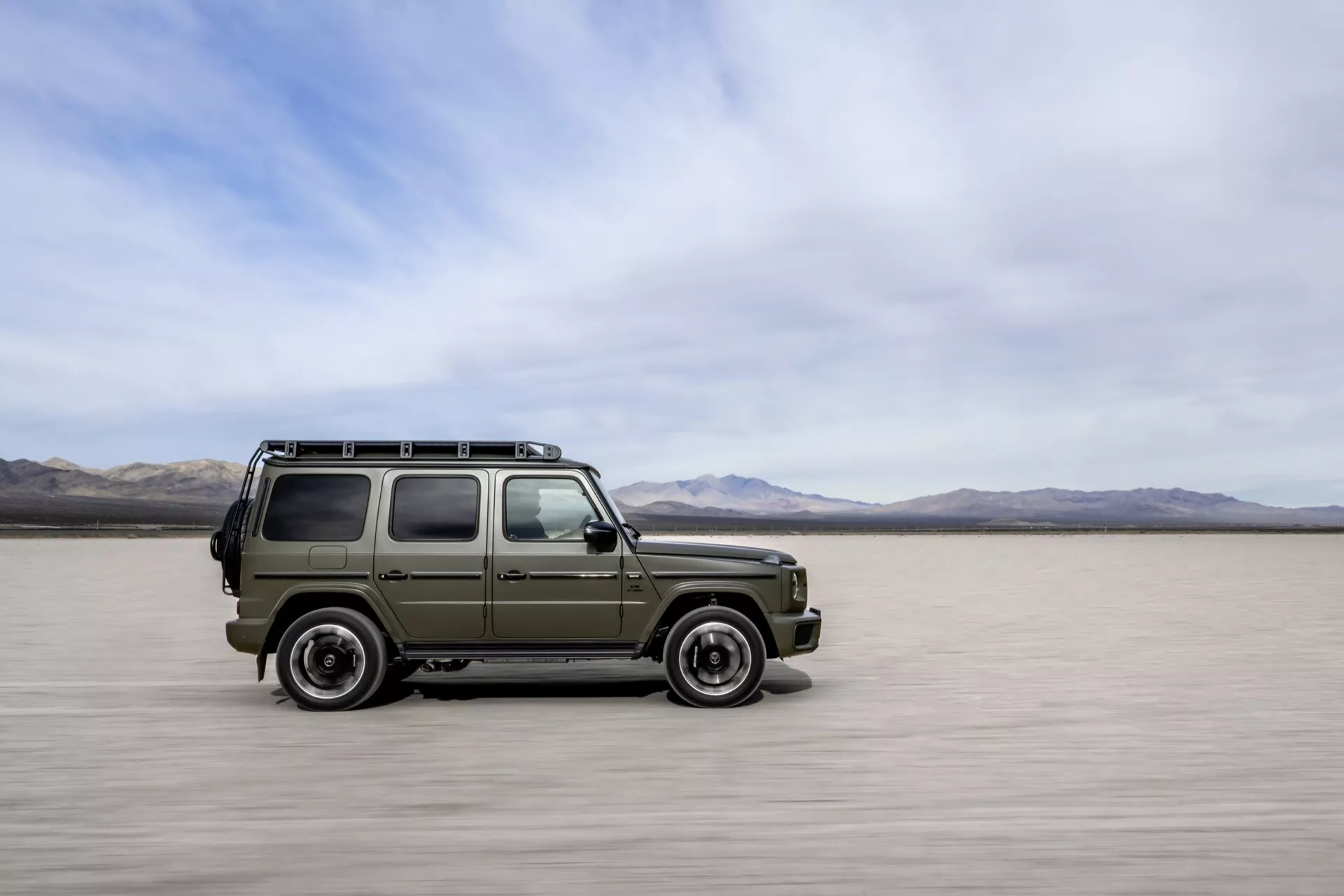 Mercedes-Benz G-Klasse 2024 v štýle vojenského SUV pohybujúci sa po solných pláňach, zobrazujúci bočný a predný profil s rozostrením v pohybe, zdôrazňujúci jeho robustný dizajn, schopnosť jazdy v teréne a tradičné prvky bez viditeľnej modernej technológie