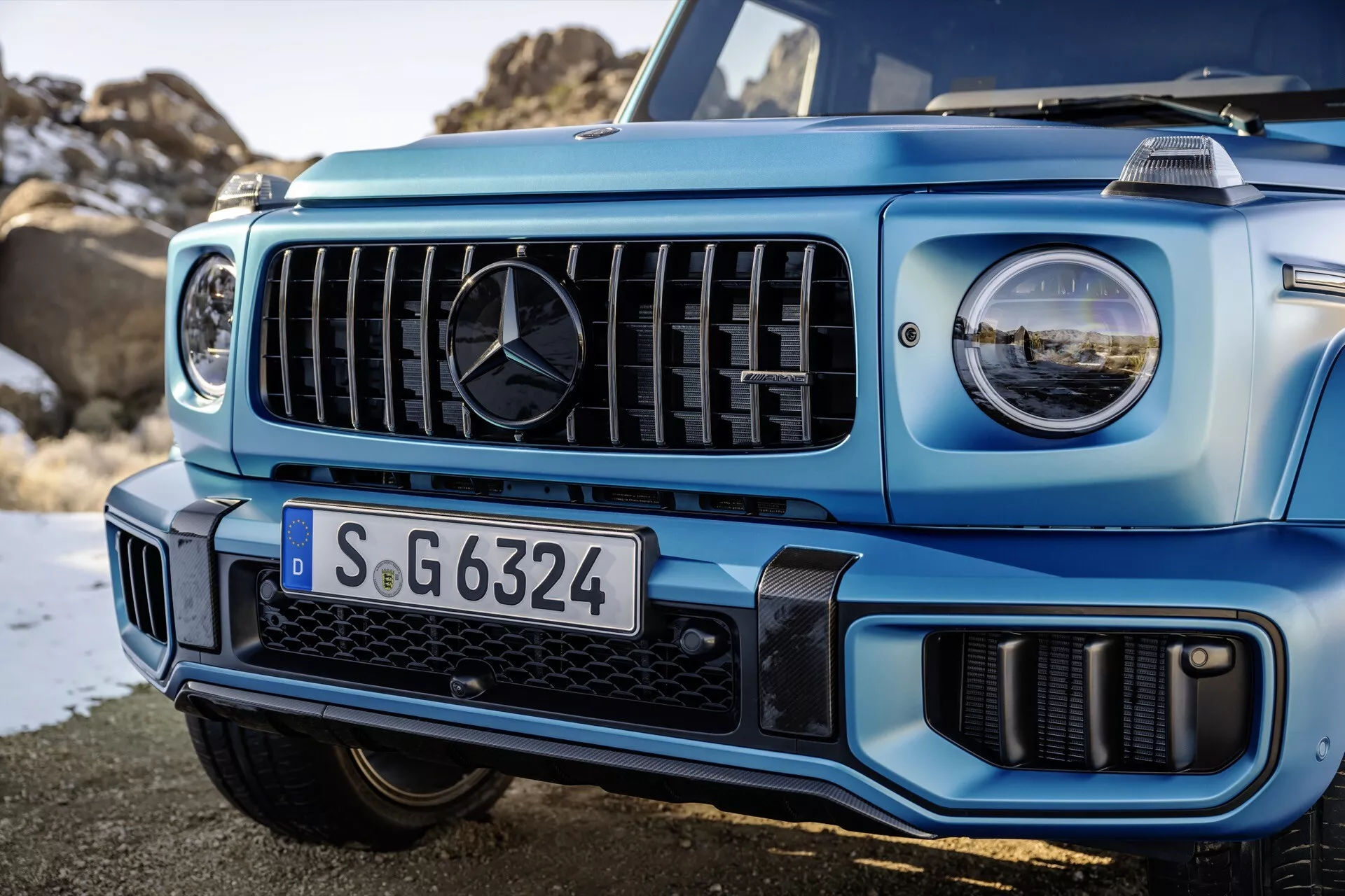Detailný pohľad na prednú ľavú stranu svetlomodrého Mercedes-Benz triedy G z roku 2024, zobrazujúci mriežku so zvislými lamelami a LED svetlomety, ilustrujúci pokročilú technológiu osvetlenia a off-road schopnosti vozidla