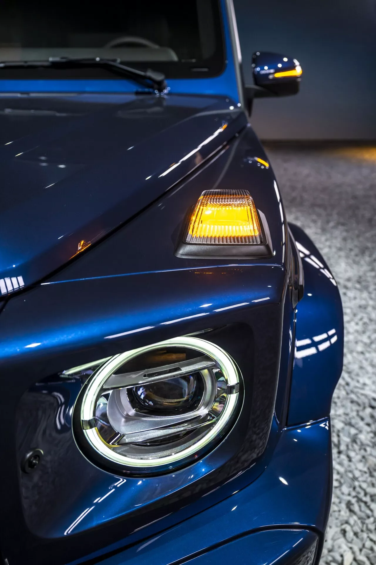 Detailný pohľad na prednú ľavú štvrtinu Mercedes-Benz G-Klasse 2024, zobrazujúci dizajn LED svetlometu, hlbokú metalízovanú modrú prémiovú úpravu a pokročilé bezpečnostné funkcie