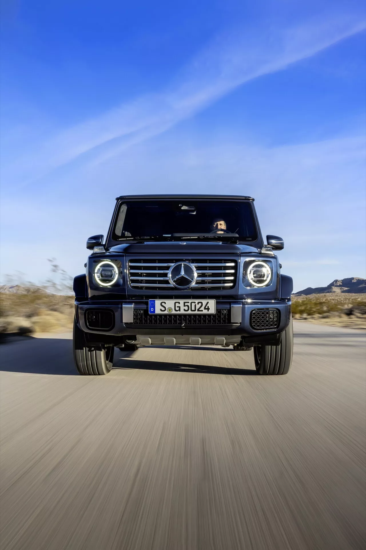 SUV Mercedes-Benz Trieda G 2024 v pohybe, zachytené z nízkeho čelného uhla, ktoré ukazuje jeho agresívny dizajn a pokročilé vlastnosti ako charakteristická mriežka, kruhové svetlomety, veľké vzduchové vstupy a zliatinové kolesá na pozadí púště