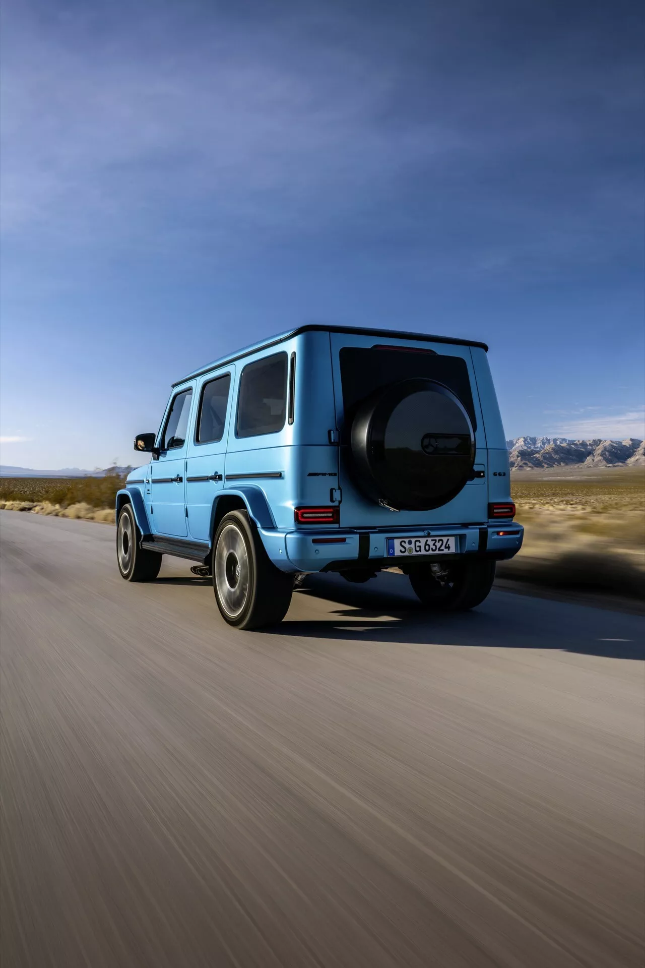 SUV Mercedes-Benz triedy G 2024 v jasnej modrej farbe zachytené z zadného trojštvrtečného uhla zobrazujúce pravý profil, zadné koleso, zadné dvere pre cestujúcich, časť strechy a namontovanú rezervnú pneumatiku na pozadí scenérie s čistou oblohou a horami, naznačujúce dobrodružstvo a výkon