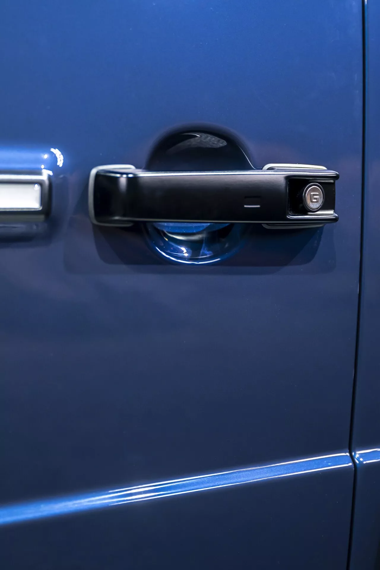Detailný záber na modrú kľučku dverí Mercedes-Benz G-Klasse 2024 s tradičným dizajnom, kľúčovou dierkou a indikátorom zamknutia, záber z boku s malou hĺbkou ostrosti