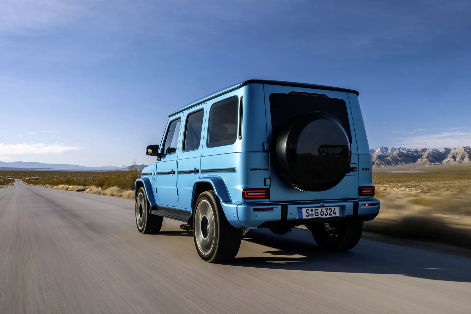SUV Mercedes-Benz triedy G ročník 2024 vo vibrantnej modrej farbe jazdiaca po spevnenej ceste s polosuchou horskou krajinou a čistým nebom na pozadí, záber z nízkeho až stredného uhla zdôrazňujúci zadnú a čiastočne stranu vodiča vozidla, typický robustný a hranatý dizajn, viditeľná rezerva na zadných dverách, výrazné blatníky a vysoká svetlá výška, kolesá s viacerými drážkami a viditeľnými brzdovými strmienkami, zadné okná chrániace súkromie, LED zadné svetlá a zadný nárazník integrovaný do farby karosérie