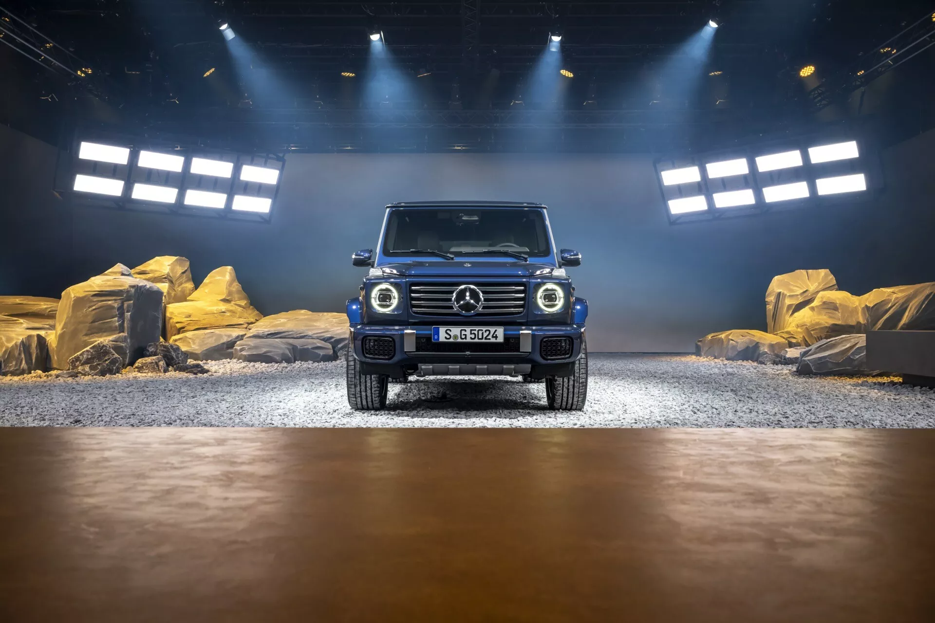 Mercedes-Benz triedy G 2024 prezentovaný v simulovanom interiéri pre jazdu v teréne, pohľad z predného uhla zdôrazňujúci mriežku chladiča, LED svetlomety a dizajn predného nárazníka, s efektmi štúdiového a prirodzeného osvetlenia zdôrazňujúcimi jeho metalický lak a schopnosti v teréne