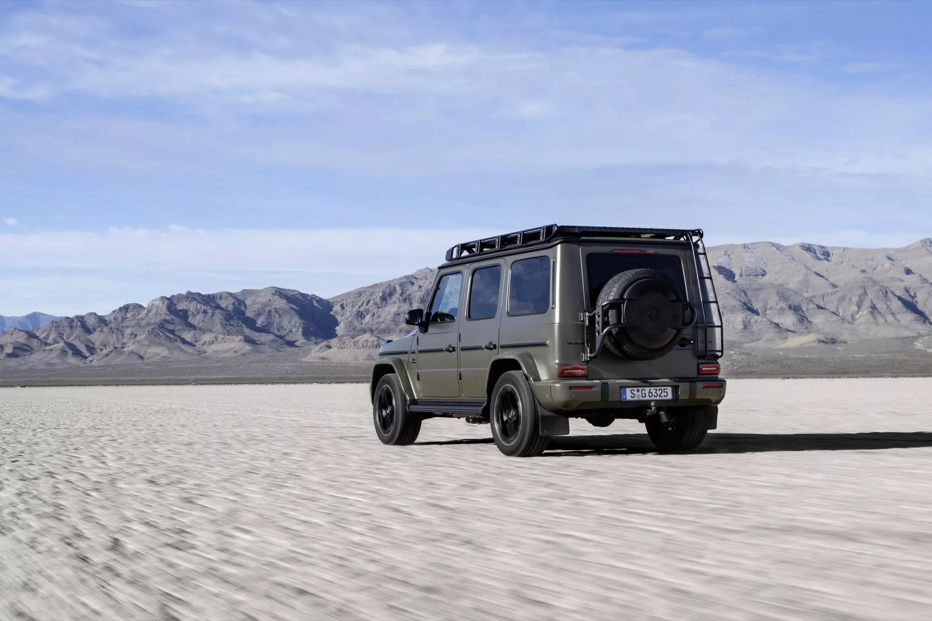 SUV Mercedes-Benz G-Klasse 2024 zachytáva jeho bočný profil s náznakom zadnej časti, predvádzajúci schopnosti pre jazdu v teréne na púštnej ploche, s nosnými koľajnicami na streche, robustnými pneumatikami a rezervným kolesom umiestneným na zadných dverách