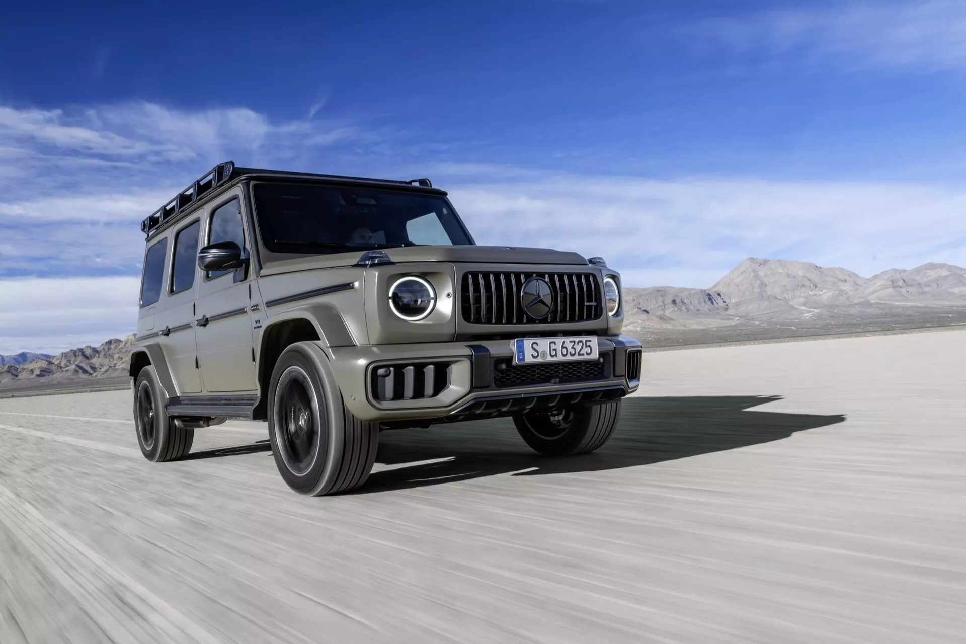 Strieborné SUV Mercedes-Benz G-Class 2024, ktoré predvádza schopnosti v off-road teréne na plochom, púštne pôsobiacom teréne, s dynamickým záberom z nízkeho uhla zdôrazňujúcim jeho robustný dizajn, kruhové svetlomety, prednú mriežku a stopu prachu.