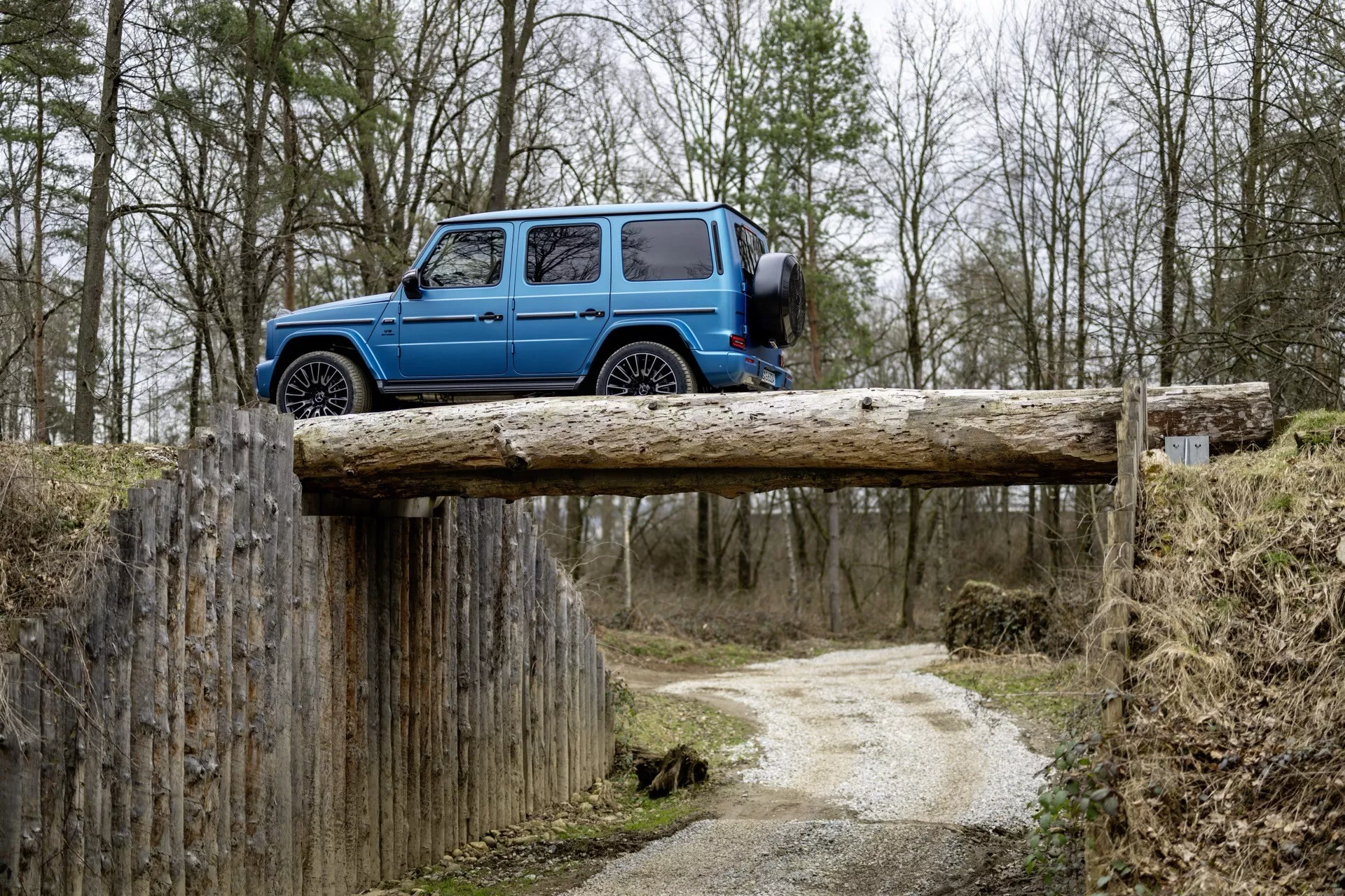 Mercedes-Benz triedy G z roku 2024 v jasne modrej farbe zaparkovaný kolmo na drevenom nosníku vo vidieckom prostredí, zdôrazňujúci jeho štvorcovú siluetu, vysokú svetlú výšku, robustné pneumatiky a funkčné dizajnové prvky, ako sú bočné stupienky, strešné okno a strešné nosiče na pozadí zamračeného lesa
