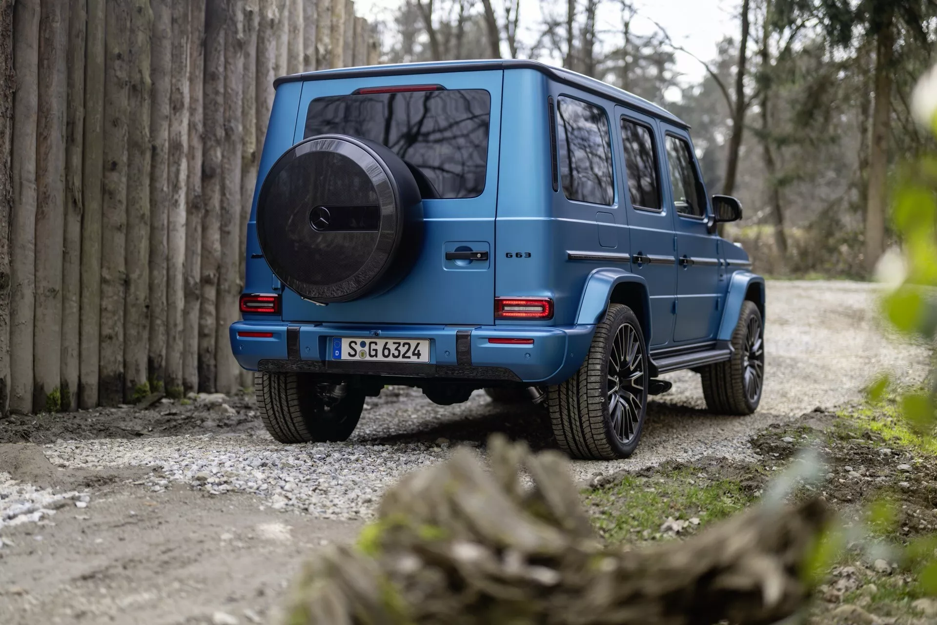 Zadný pohľad na modré SUV Mercedes-Benz triedy G (G 465) 2024, zdôrazňujúci jeho robustný, škatuľový dizajn, rezervné koleso na zadných dverách, LED zadné svetlá, veľkú svetlosť a zliatinové kolesá na štrkovej ceste s prírodným prostredím