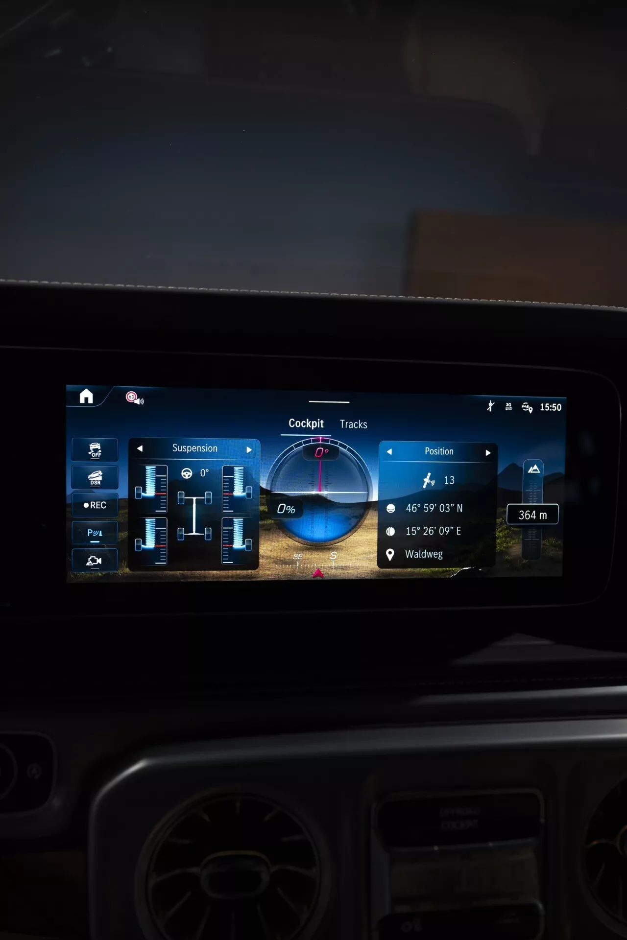 Detailný pohľad na moderný dotykový displej informačno-zábavného systému v Mercedese-Benz G-Klasse 2024 s navigáciou, nastavením zavesenia, terénnymi trasami pre jazdu v teréne a ovládaním multimédií, odrážajúci pokročilé technológie automobilov a luxusný dizajn interiéru.
