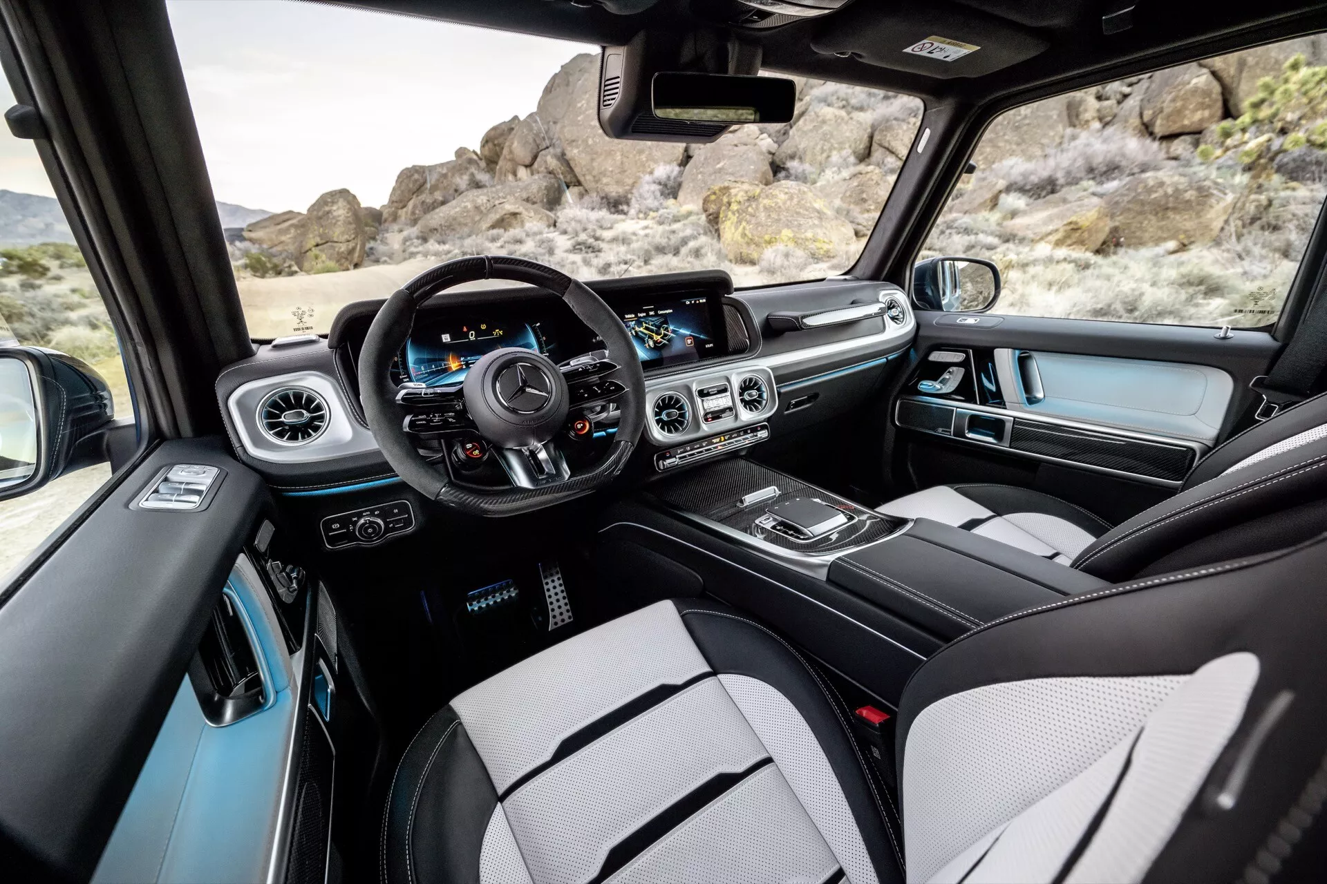 Interiér Mercedes-Benz G-Klasse 2024 so zameraním dopredu zo zadnej sedadlovej časti, zobrazujúci centrálny a vodičovú stranu palubnej dosky, ovládacie prvky na dverách vodiča, volant s multifunkčnými tlačidlami, panel prístrojov, systém infotainment, ovládanie klímy, centrálnu konzolu a čiastočný výhľad na strešné okno.