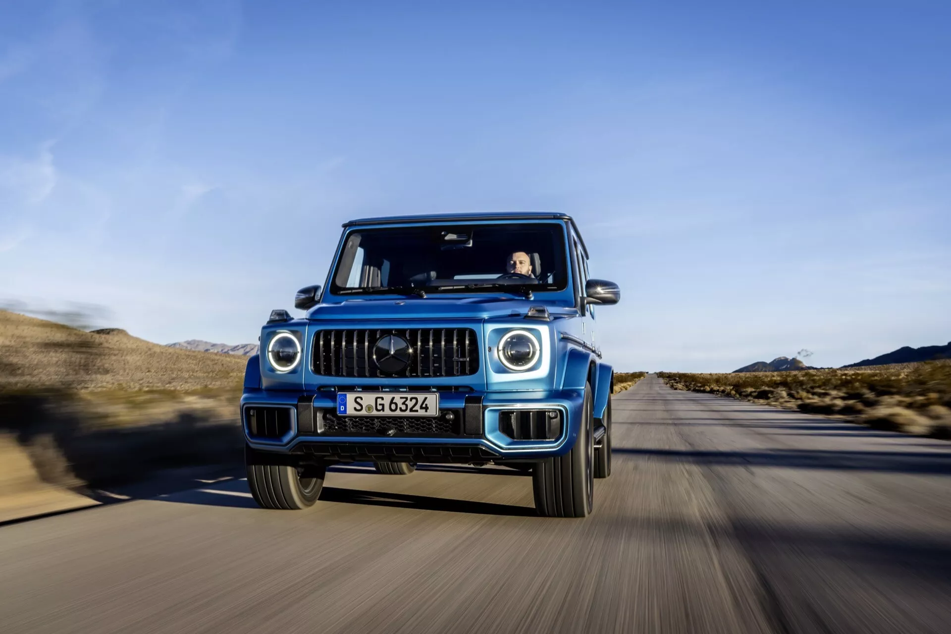 Luxusné SUV Mercedes-Benz triedy G 2024 v pohybe, zachytené z prednej perspektívy s technikou sledovacieho záberu zobrazujúce detailnú mriežku, LED svetlomety a funkcie ADAS, na púštnej ceste pod čistou modrou oblohou