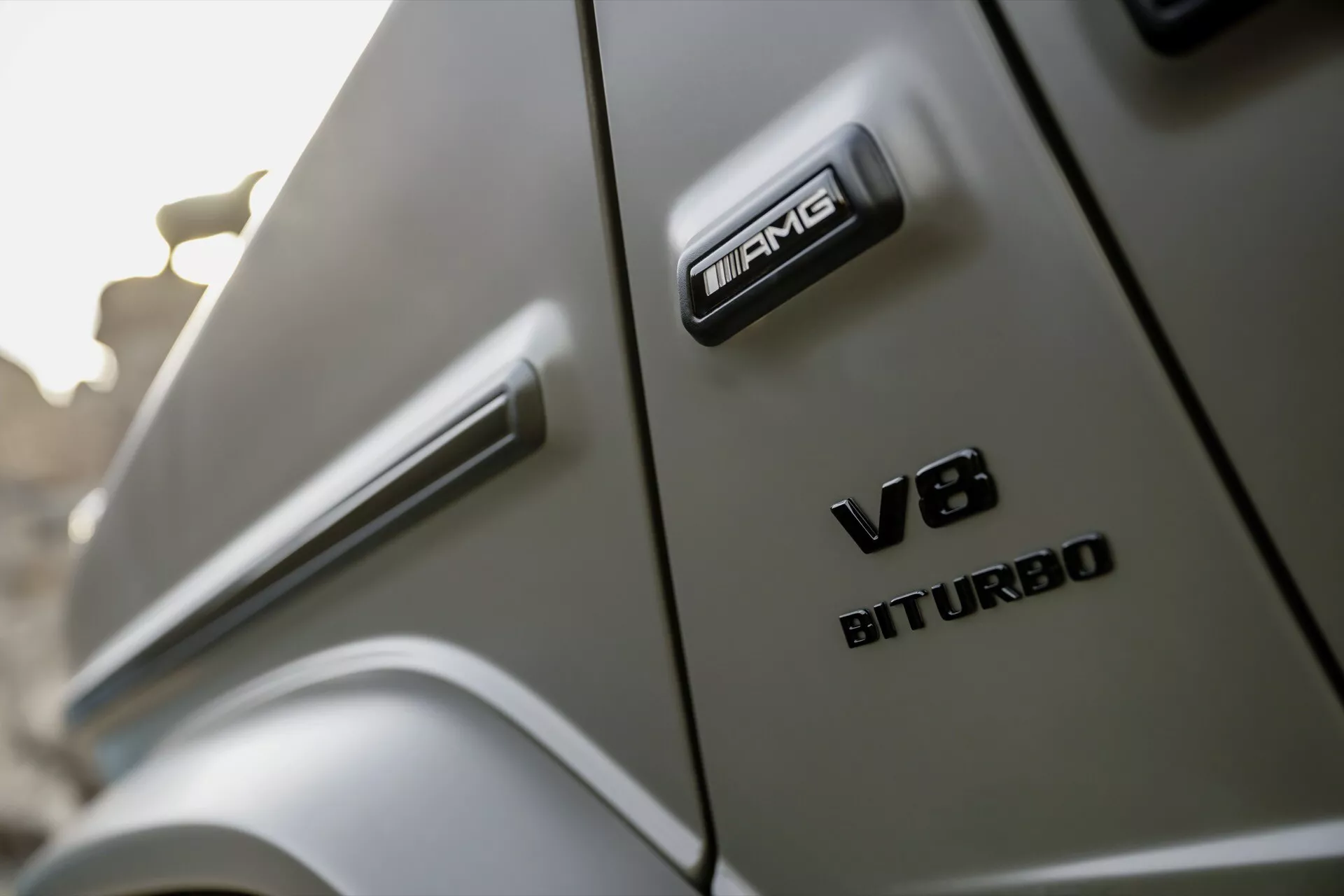 Detailný pohľad z uhla zobrazujúci odznaky AMG a V8 BITURBO na novom Mercedes-Benz G-Klasse 2024, zdôrazňuje prvky vysokého výkonu a luxusného dizajnu vozidla.