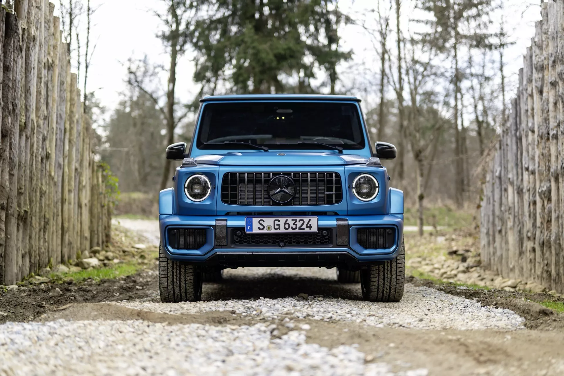 Čelný pohľad na modré SUV Mercedes-Benz G-Klasse 2024 na štrkovej ceste, medzi radmi drevených stĺpikov, zdôrazňujúci moderný dizajn vozidla a LED svetlomety.