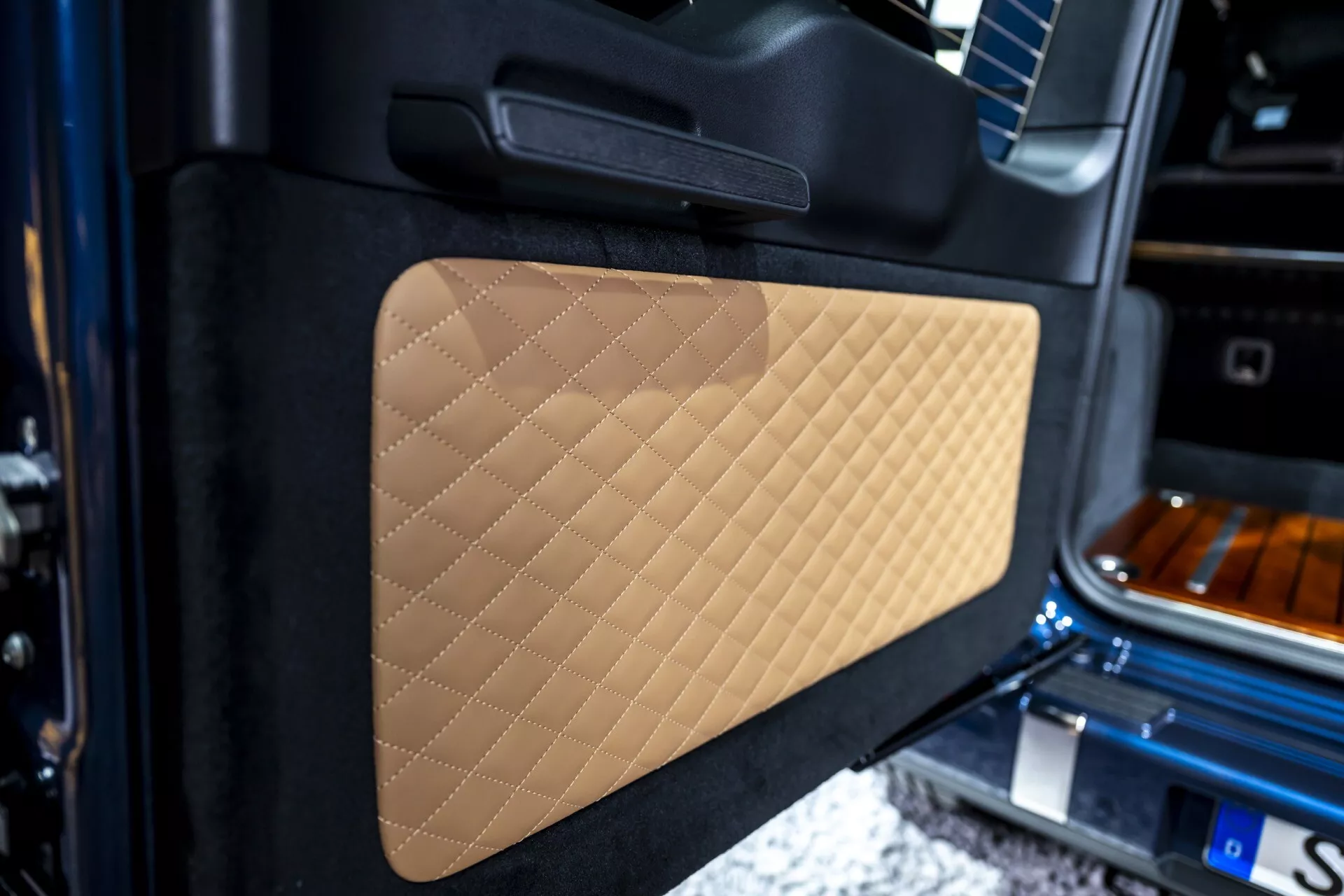 Detailný pohľad na vnútorný panel dverí Mercedes-Benz triedy G (G 465) na rok 2024 s prešívaným béžovým koženým detailom a čiernou rukoväťou, zdôrazňujúci luxusné remeselné spracovanie a dizajn