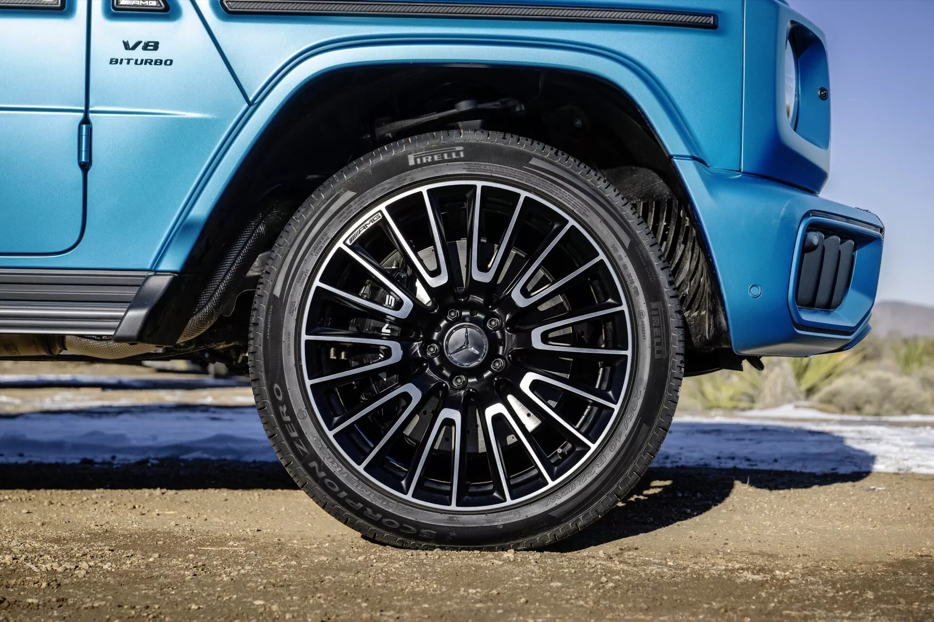 Mercedes-Benz triedy G (G 465) pre rok 2024 predvádza zadné koleso na strane spolujazdca s viacramenným dizajnom a tmavou povrchovou úpravou na pozadí živé modrej karosérie, vrátane detailu na emblému V8 BITURBO a detailu bočného nástupního schodíka