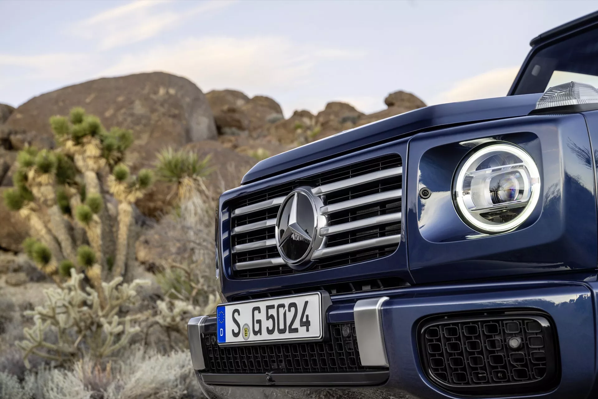 Detailný pohľad na prednú ľavú stranu Mercedes-Benz G-Klasse 2024 s výraznou mriežkou a centrálnym emblémom, časťou pokročilého LED svetlometu, na pozadí s prírodným exteriérom naznačujúcim schopnosti v teréne