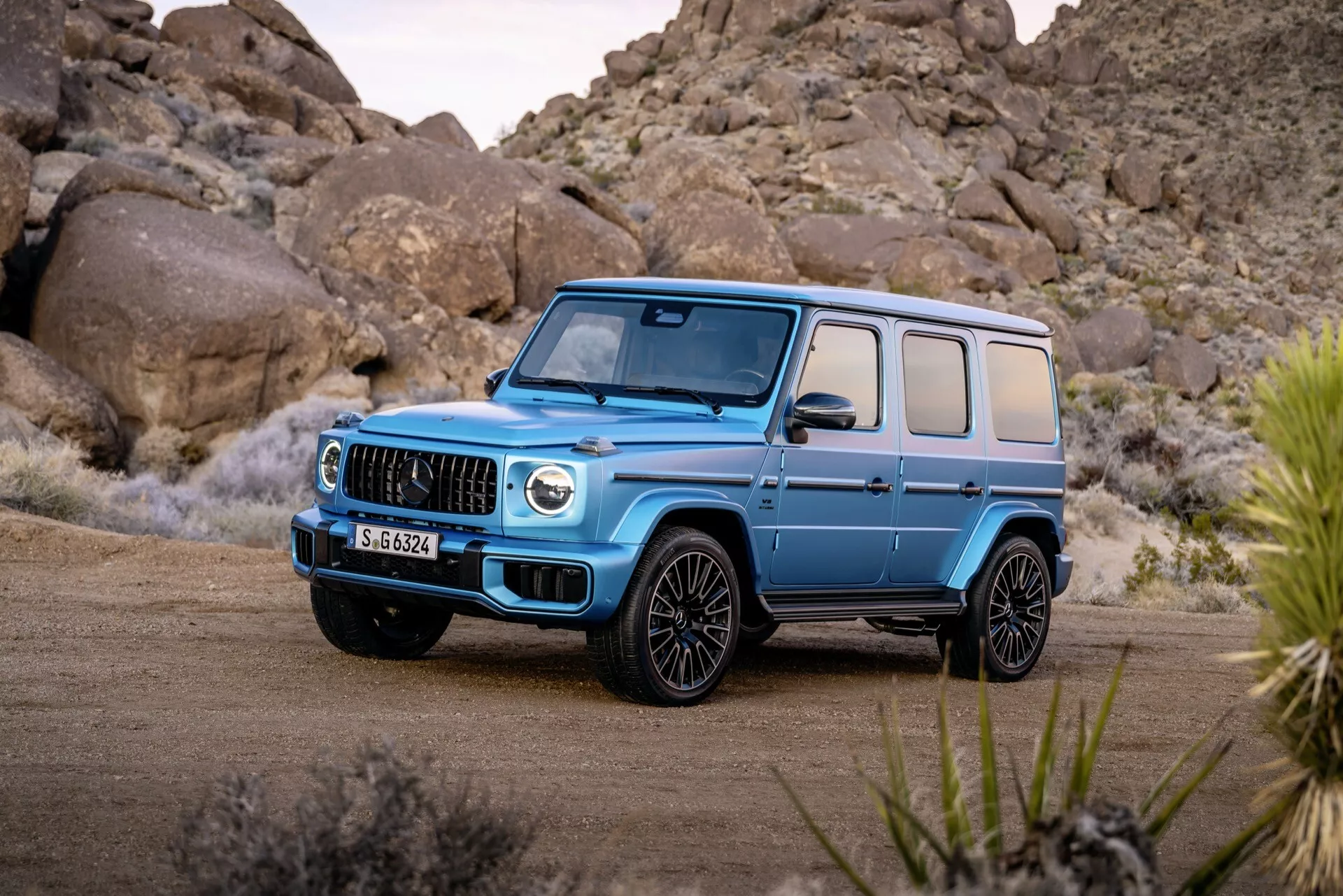 Luxusné SUV Mercedes-Benz G-Klasse 2024 v svetlomodrej farbe, predstavuje jeho charakteristickú mriežku, veľké vstupné otvory a LED svetlomety z uhla prednej časti a boku na nezpevnenej ploche so skalnatou púšťou na pozadí, čo zdôrazňuje jeho schopnosti v teréne a moderné konštrukčné prvky.
