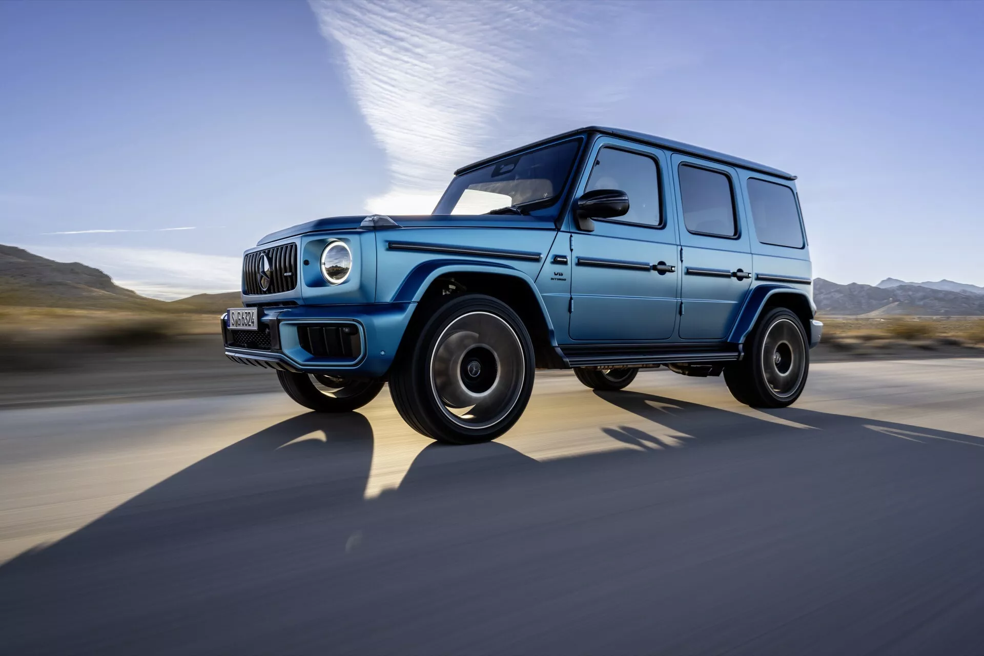 Luxusné SUV Mercedes-Benz triedy G roku 2024 zachytené v pohybe z dynamickej nízko-uholnej bočnej pohľadu, ktoré zvýrazňuje jeho škatuľovitý, robustný dizajn, modrú farbu s čiernymi prvkami, okrúhle svetlomety, LED denné svietenie a čierne zliatinové kolesá proti pozadiu rozostreného vysokou rýchlosťou.