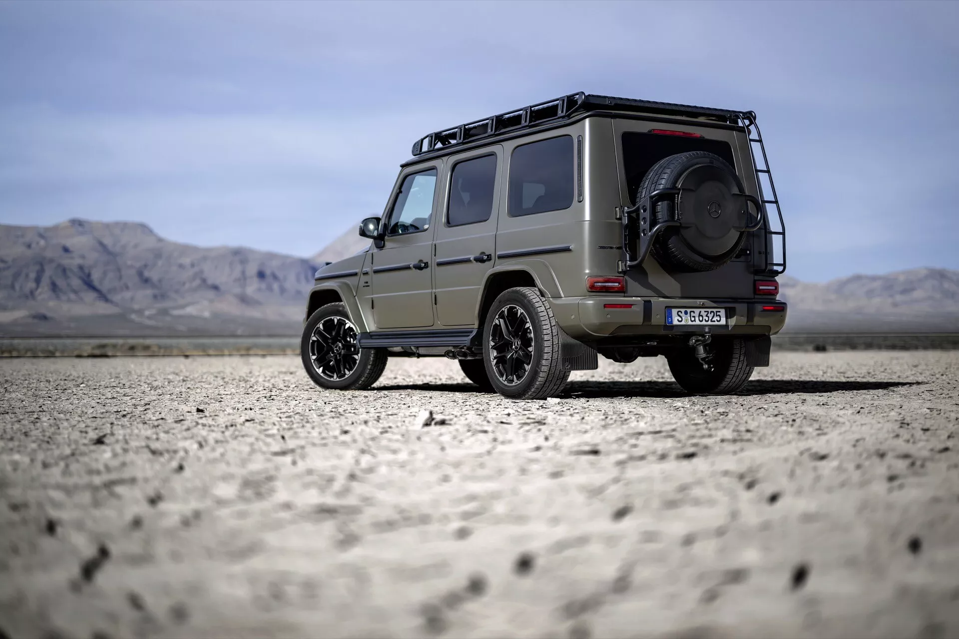 SUV Mercedes-Benz triedy G 2024 zaparkované na pustátnej jaziernej pláni, ukazujúce jeho robustný dizajn so strešným nosičom, náhradným kolesom na zadnej časti a štýlovými zliatinovými disky pod čistou oblohou s ďalekými horami.