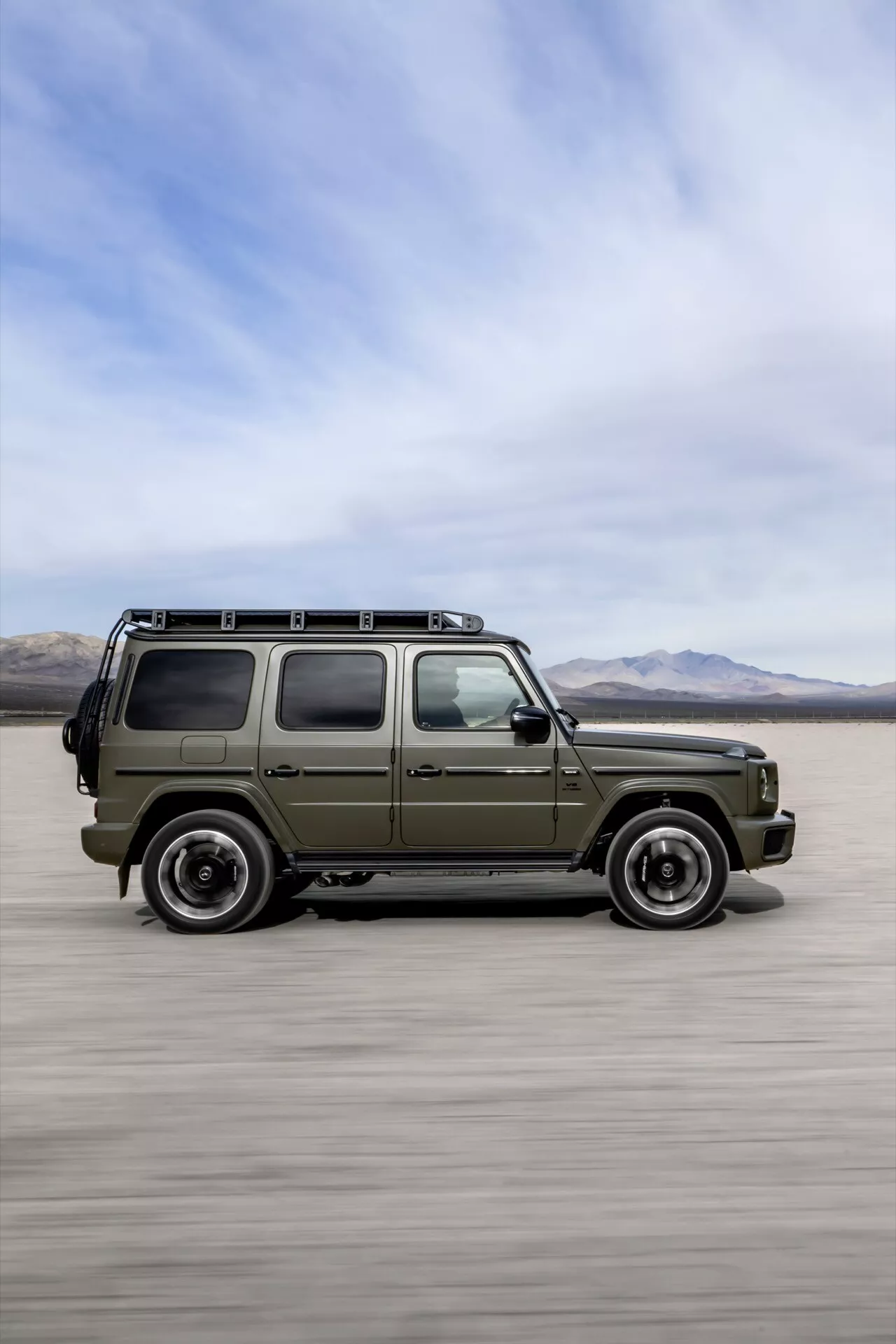 SUV Mercedes-Benz triedy G (G-Klasse 465) z roku 2024 sa pohybuje po rovnom púštňom teréne s horami v pozadí, čo zdôrazňuje jeho olivovo zelenú karosériu, bočný profil, strešný nosič a efekt rozostrenia pohybu