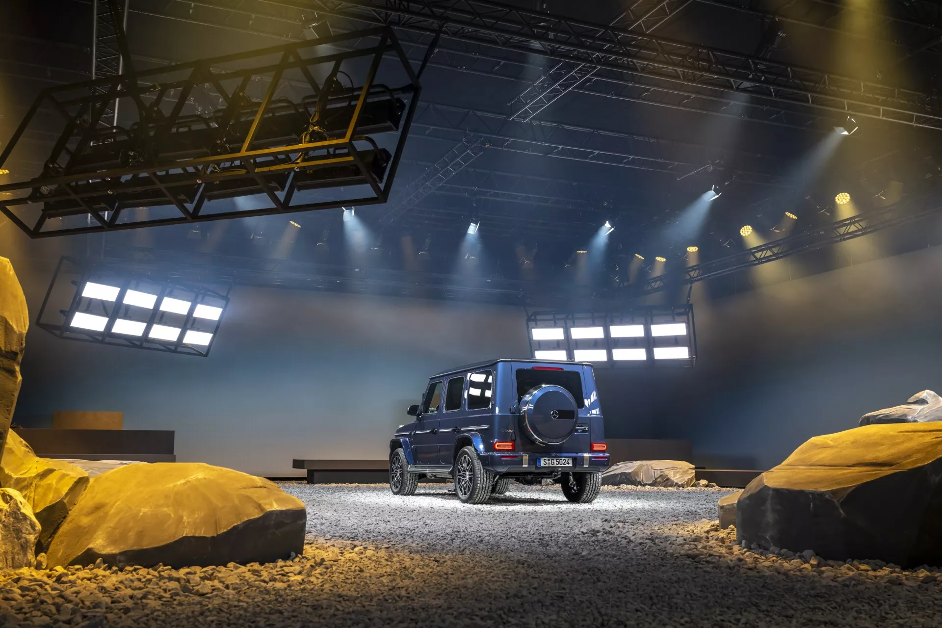Čierne SUV Mercedes-Benz Triedy G (G-Klasse 465) rok 2024 z pohľadu tri štvrtiny zozadu prezentujúce schopnosti pre jazdu v teréne s rezervným kolesom, LED zadnými svetlami a luxusným dizajnom v inscenovanom terénnom simulovanom vnútornom prostredí s štrkom a štruktúrami pripomínajúcimi skaly pod dramatickým umelým osvetlením