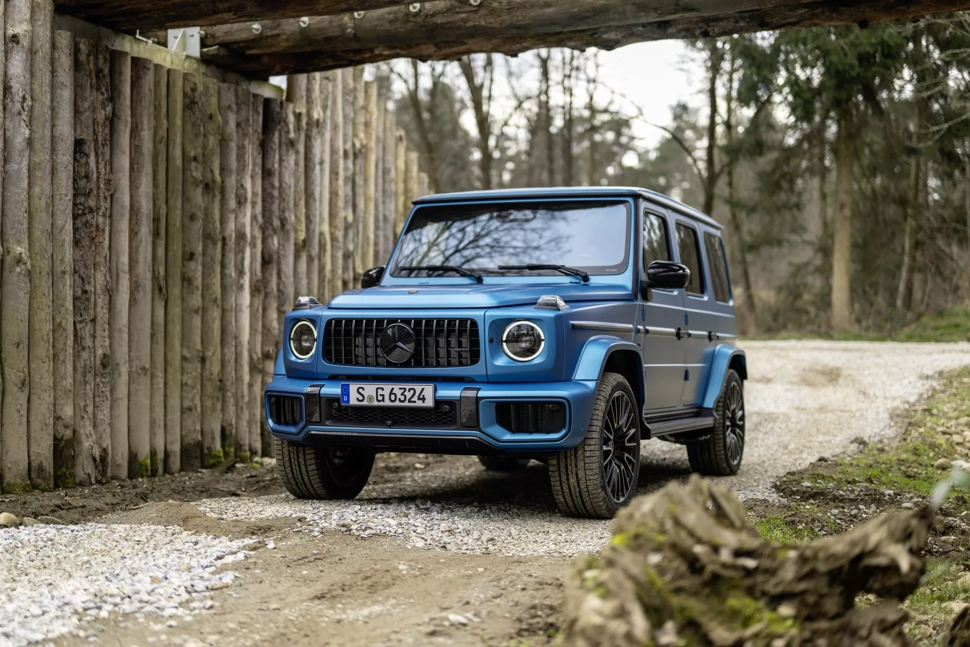 Trojštvrťový predný pohľad na modré SUV Mercedes-Benz G-Class 2024, zdôrazňujúci prednú mriežku, LED svetlomety, predný nárazník a zliatinové kolesá, na pozadí rustikálneho terénu so soft osvetlením.