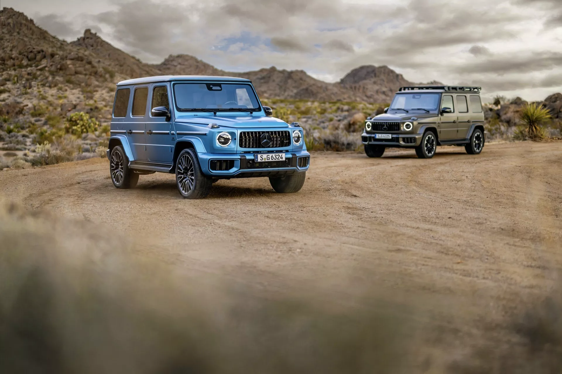 Mercedes-Benz triedy G 2024 v svetlomodrej farbe, dôrazne zobrazený v púštnej krajine na prašnej ceste, prezentujúci LED svetlomety, vertikálne chrómové mriežky, veľké disky z ľahkej zliatiny s viacerými ramenami a pneumatiky vhodné do terénu, a pokročilé technologické funkcie, so druhým SUV triedy G v olivovej zelenej farbe viditeľným na pozadí.