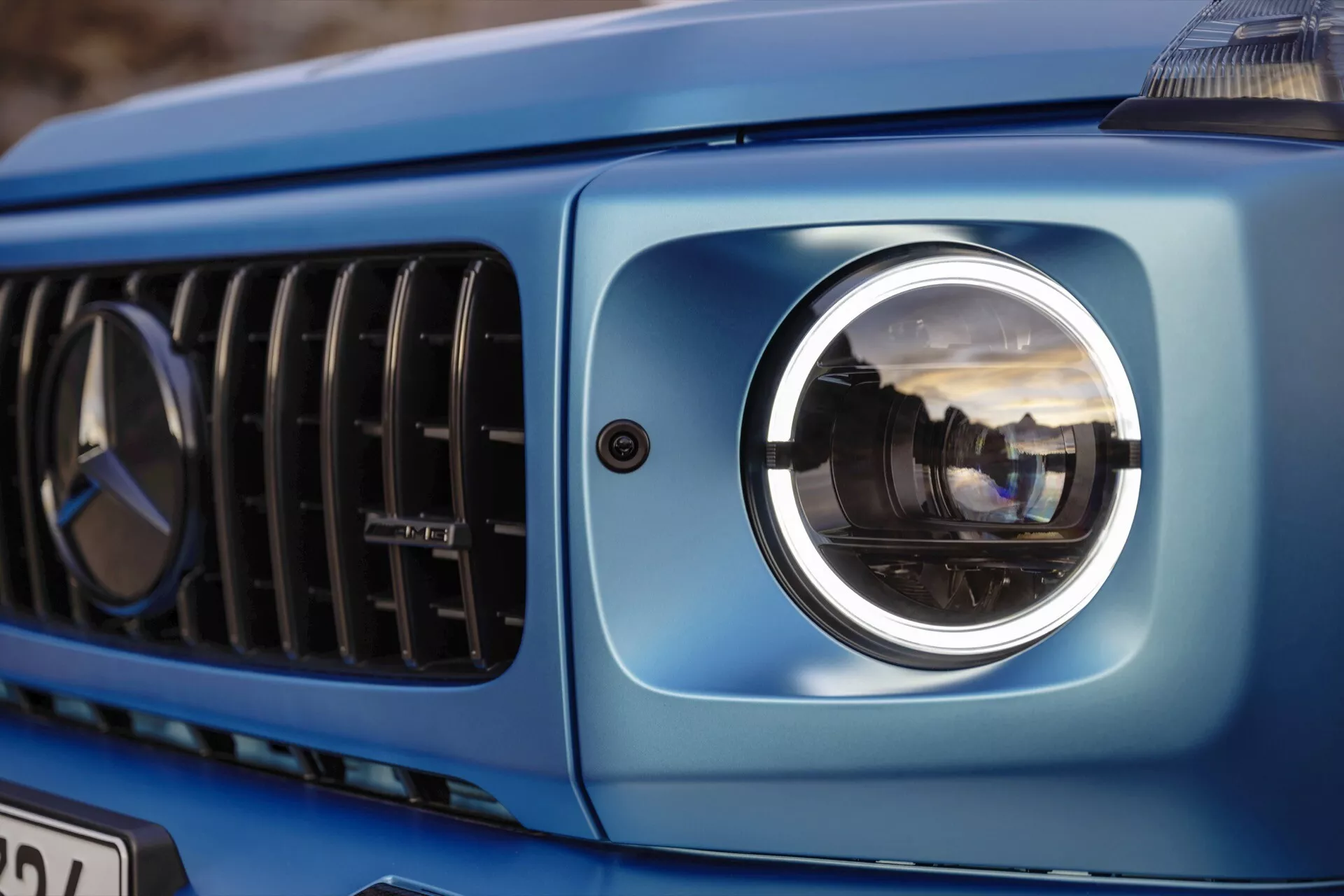 Detailný pohľad na prednú ľavú časť Mercedes-Benz triedy G 2024, zdôrazňujúci moderný projektorový svetlomet, časť mriežky a metalické modré karosérie