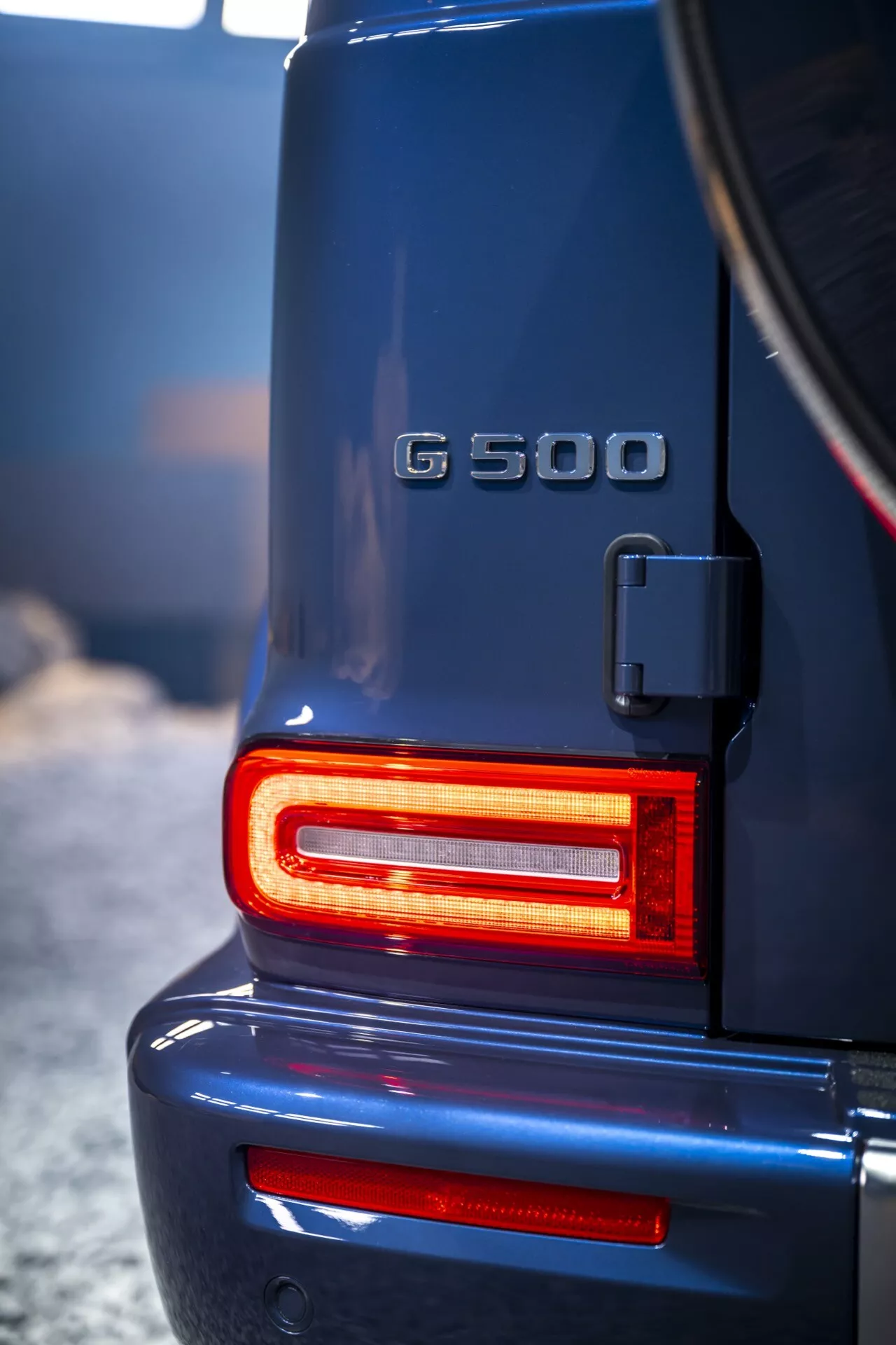 Zadná časť Mercedes-Benz triedy G (G 500) rok 2024 s modernými LED zadnými svetlami svietiacimi na červeno, ktoré zdôrazňujú vysokokvalitný, robustný dizajn a symbol statusu luxusného SUV s jasným pohľadom na nosič rezervného kolesa a označenie modelu.