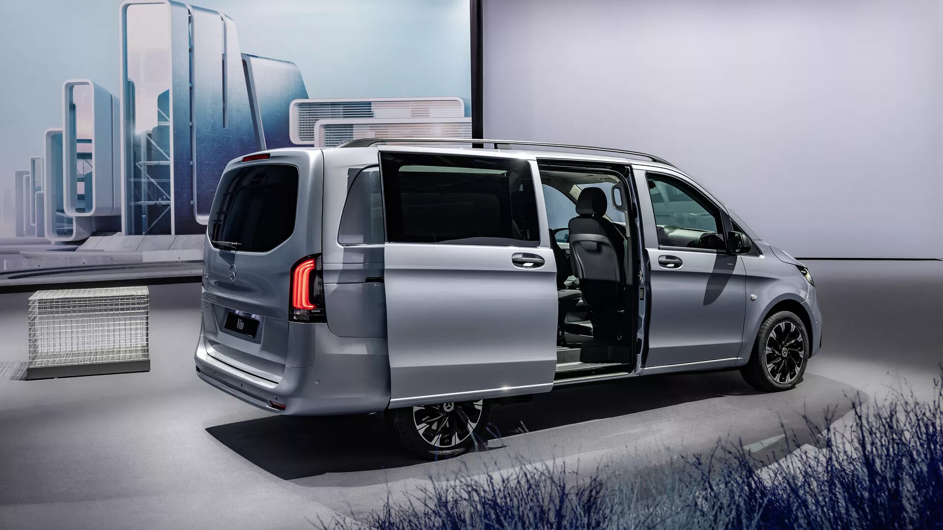 Strieborný minivan Mercedes-Benz Vito 2024 (447 facelift) s otvorenými posuvnými bočnými dverami ukazujúci elegantný moderný dizajn z trojštvrťového zadného uhla, zdôrazňujúci aerodynamický profil vozidla, zadné svetlá LED, zliatinové kolesá a náhľad do pokročilého interiéru s potenciálne prémiovým materiálom sedadiel a modernou palubnou doskou.