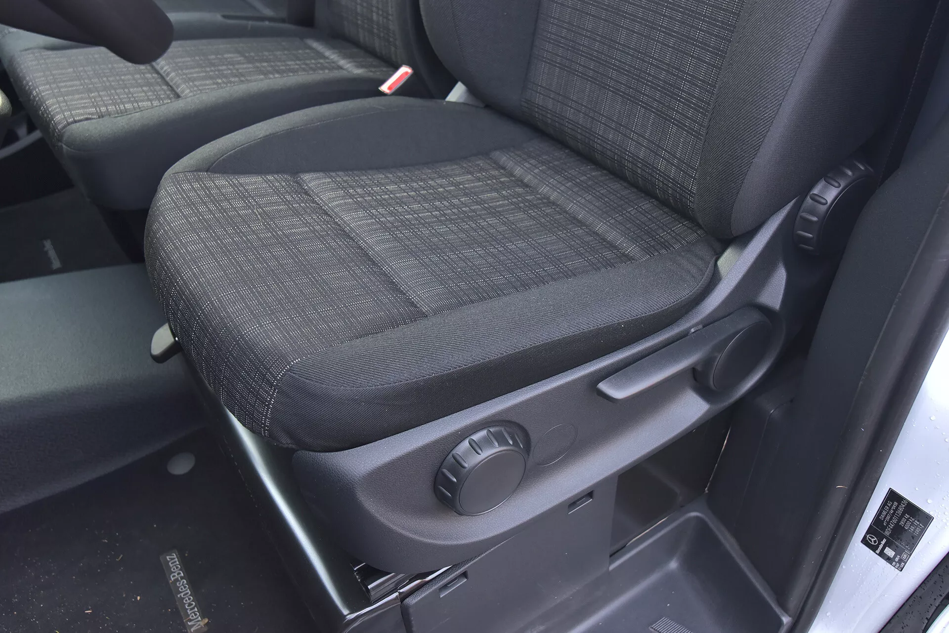 Interiér Mercedes-Benz Vito 2014 Tourer so zameraním na spolujazdcove sedadlo s odolným tmavým poťahom, ovládacím gombíkom pre manuálne nastavenie a bezpečnostnými prvkami ako ľahko viditeľný prijímač bezpečnostného pásu.