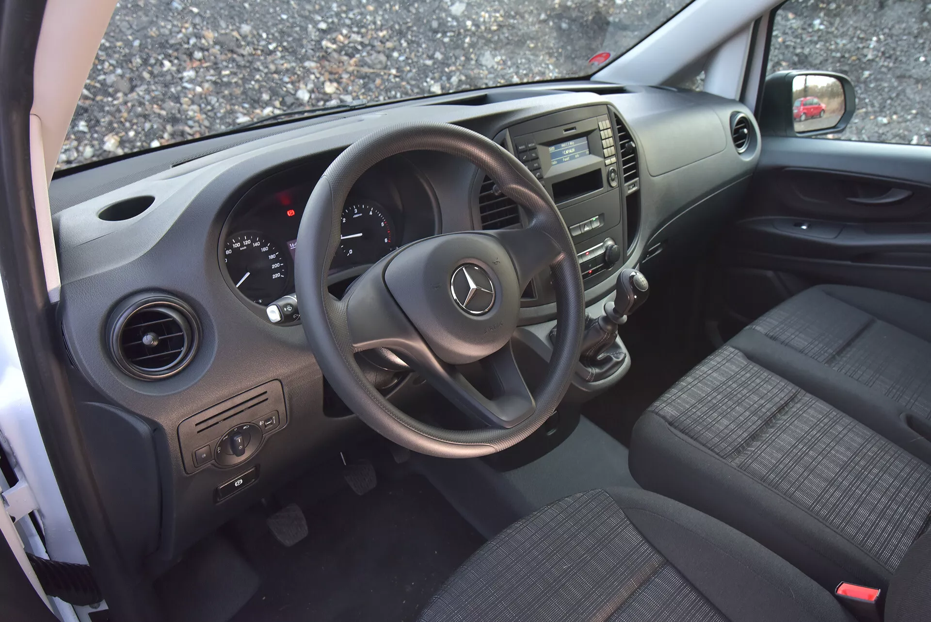 Interiér Mercedes-Benz Vito Tourer z roku 2014 z pohľadu z otvorených dverí vodiča, zvýrazňujúci volant s logom Mercedes, prístrojový panel, stredovú konzolu, sedadlá z látky, ovládacie prvky na dverách a ventilačné otvory.