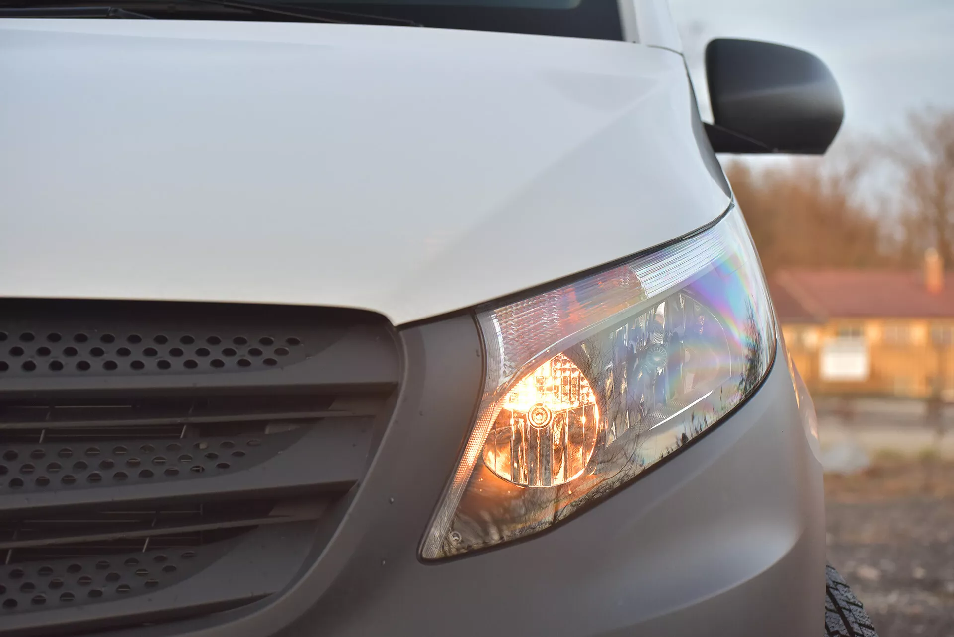 Zbližný bočný pohľad na biely Mercedes-Benz Vito Tourer z roku 2014 zameraný na svietiaci predný svetlomet, časť mriežky a kapotu, zdôrazňujúci jeho halogénové svetlomety a aerodynamický dizajn v mäkkom osvetlení.