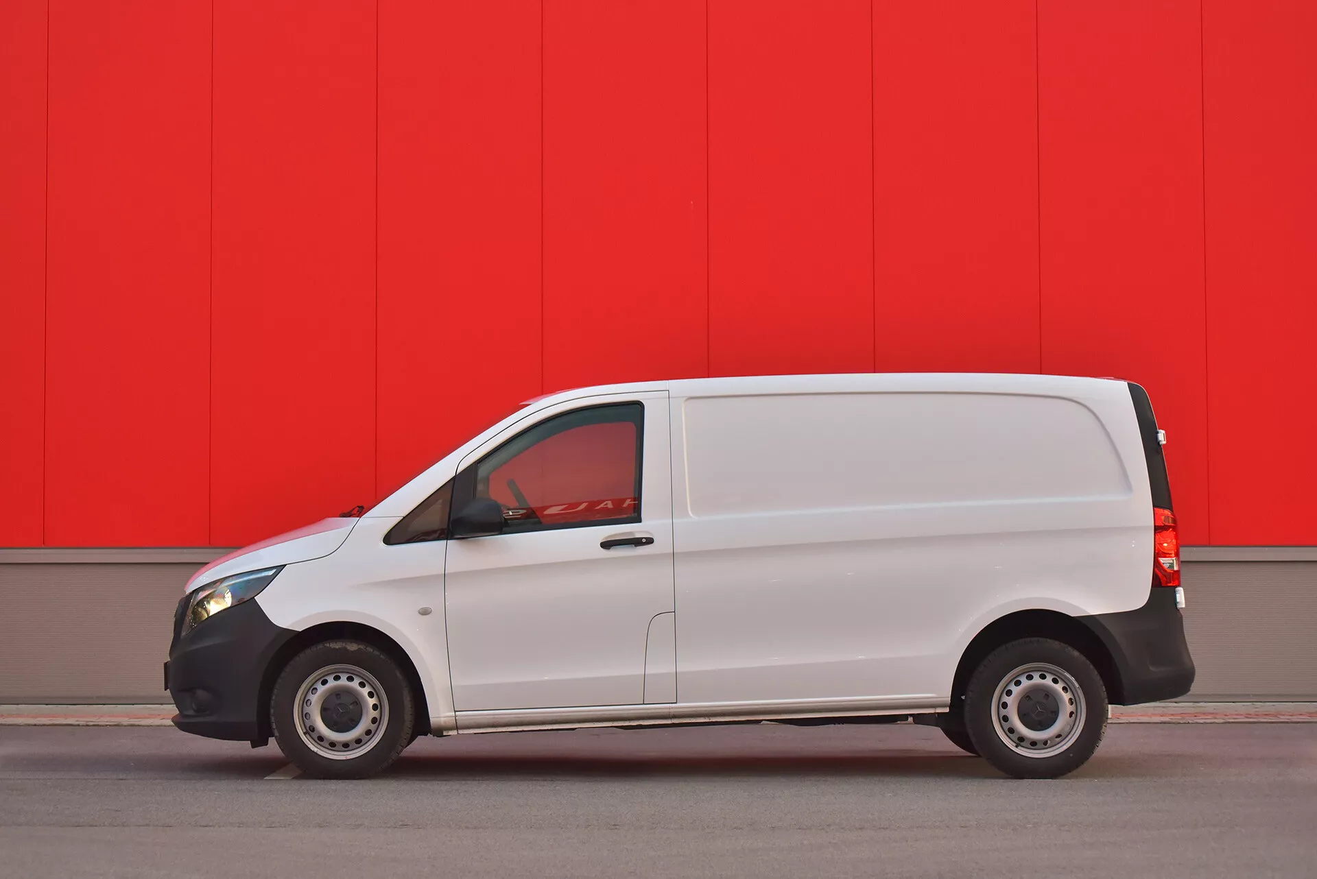 Biela dodávka Mercedes-Benz Vito Tourer z roku 2014 zaparkovaná paralelne proti žiarivo červenej stene, zobrazuje bočný profil s viditeľnými dverami, oknami a oceľovými kolesami, zachytená z nízkeho uhla na zdôraznenie dizajnu a funkcií vozidla