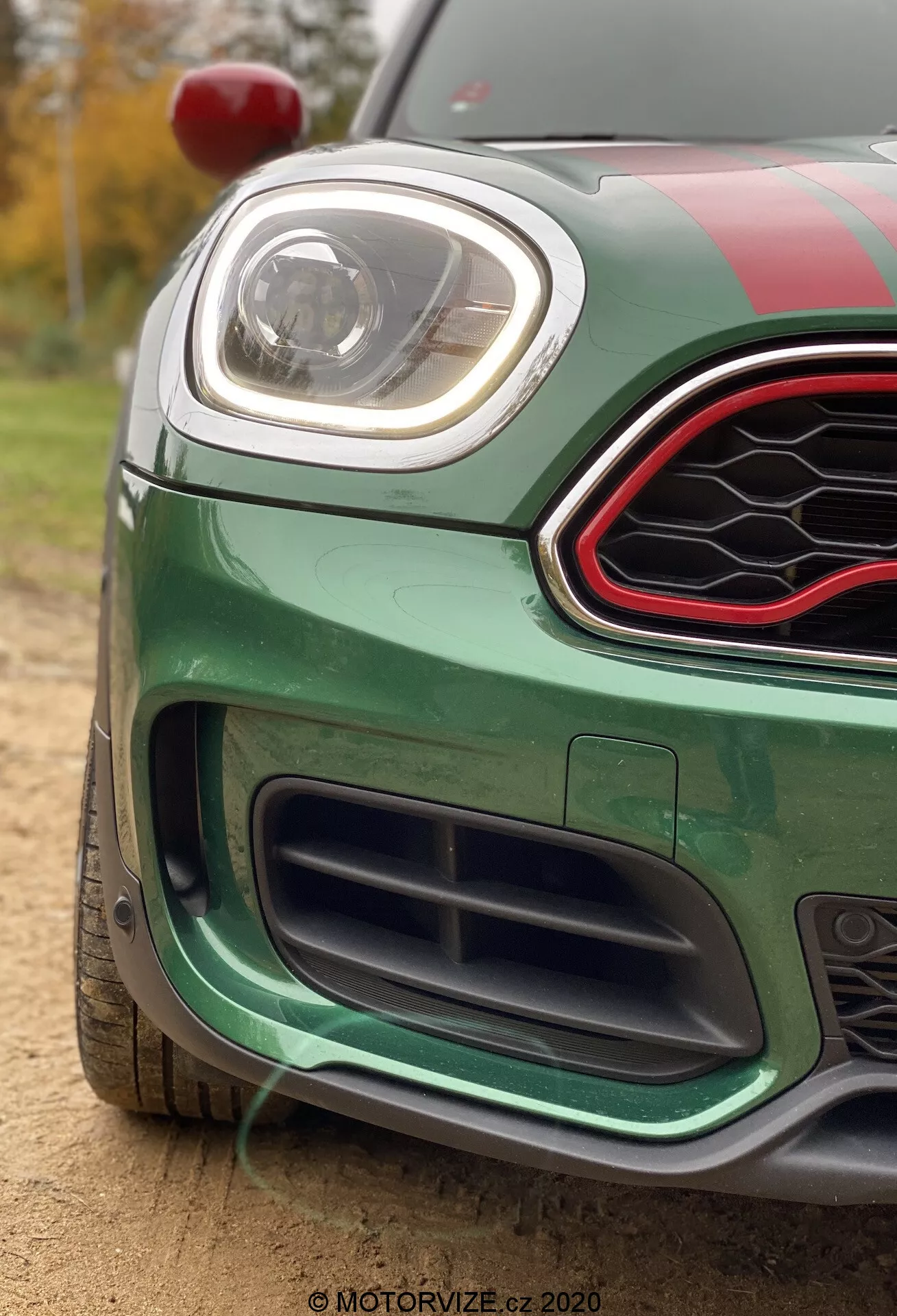 Záber na zelené Mini Countryman John Cooper Works 2020, zameraný na ľavú prednú štvrtinu. Fotografia je urobená z nízkeho uhla, čo podčiarkuje dolnú mriežku s čiernym voštinovým dizajnom, predný nárazník s aerodynamickými prvkami a elegantný LED svetlomet. Horná mriežka má šesťuholníkový tvar s lesklou čiernou povrchovou úpravou a červeným akcentným lemovaním. Auto má závodné pruhy na kapote a streche, červené bočné zrkadlo viditeľné v ľavom hornom rohu a je umiestnené vonku za denného svetla s rozmazaným pozadím obsahujúcim stromy a zeleň.