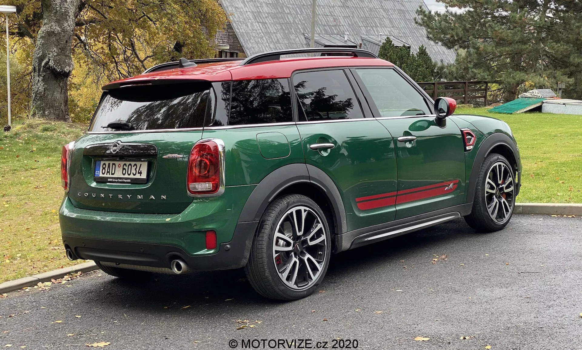Trojštvrtinový zadný pohľad na zelený MINI Countryman John Cooper Works z roku 2020, zaparkovaný na obytnom príjazdovom chodníku. Obrázok ukazuje celú zadnú polovicu pravej strany auta, vrátane zadného výklopného dvere, pravého zadného kolesa, časti strechy a dverí pre pasažierov. Športový a kompaktný SUV dizajn obsahuje kontrastné červené akcenty na krytoch bočných zrkadiel a streche, čierne oblúky kolies a veľké viacramenné zliatinové kolesá. Zadné svetlá, názov modelu 'Countryman' a logo MINI sú výrazne viditeľné. Auto má červenú strechu so spoilerom a strešnými ližinami a zadné okno obsahuje odmrazovač a stierač.