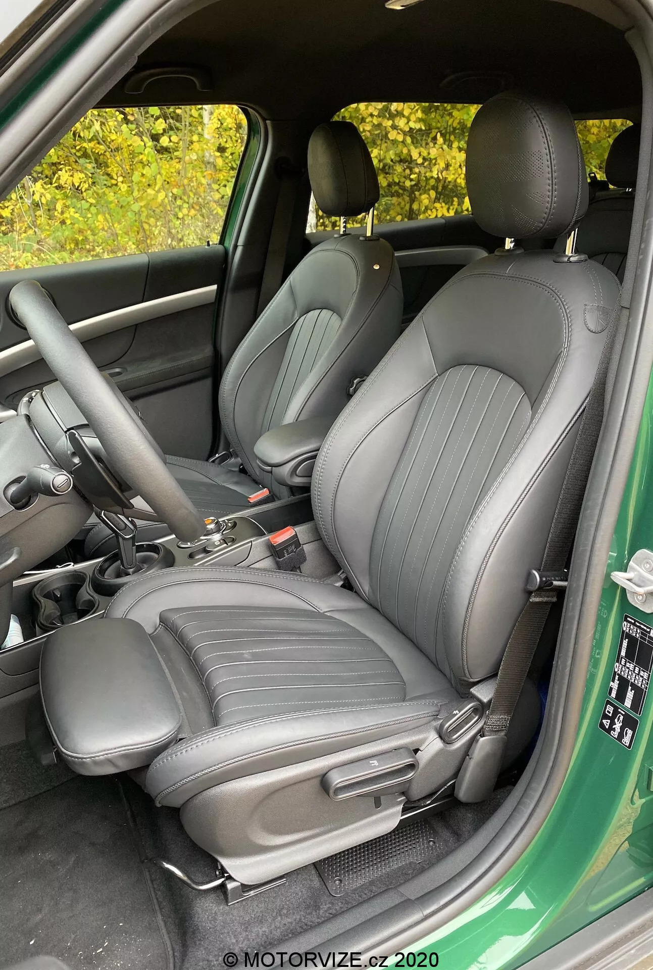 Obrázok interiéru Mini Countryman John Cooper Works 2020 zameraný na predné sedadlá vodiča a spolujazdca, čalúnené tmavým vysokokvalitným materiálom s vertikálnymi čiarami, pohľad z otvorených dverí vodiča, zobrazujúci stredovú konzolu s radiacou pákou a ručnou brzdou, čiastočný pohľad na dverný panel s ovládaním okien a zrkadiel.