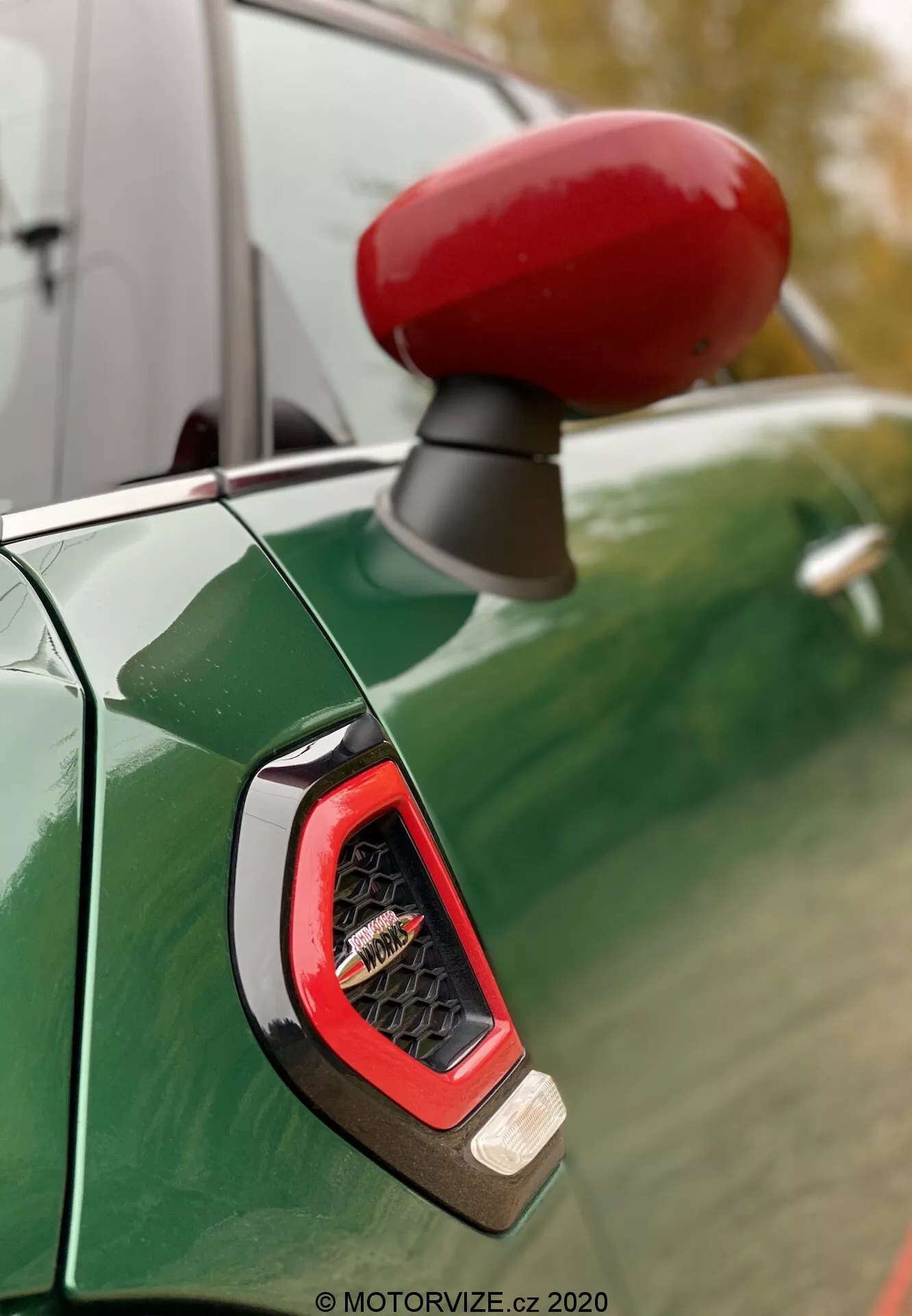 Detailný záber na svietivo červené krytie bočného zrkadla a bočný ukazovateľ na strane vodiča Mini Countryman John Cooper Works z roku 2020, viditeľný proti hlbokému metalickému zelenému karosérii s odrazmi zamračenej oblohy a stromov. Zaostrenie zdôrazňuje uhlový, číry ukazovateľ pravdepodobne využívajúci LED technológiu, obklopený športovým a lesklým povrchom.