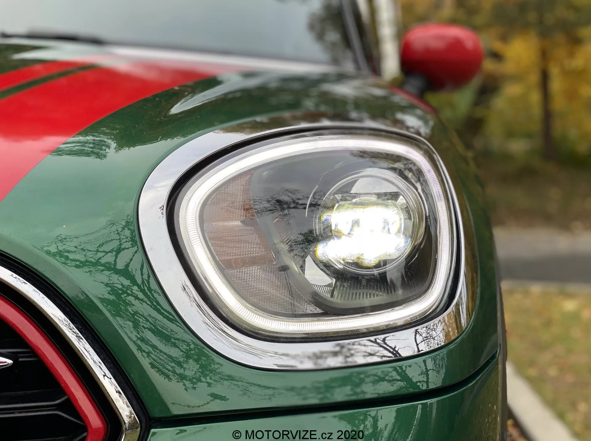 Blízky pohľad na prednú ľavú časť 2020 Mini Countryman John Cooper Works, zameraný na moderný okrúhly svetlomet s LED technológiou, kryt svetlometu a osvetlený projektorový reflektor. Obrázok zobrazuje zakrivenie predného blatníka, okraj kapoty a časť mriežky s chrómovou úpravou. Auto má lesklý, metalický zelený lak s viditeľnými metalickými vločkami.