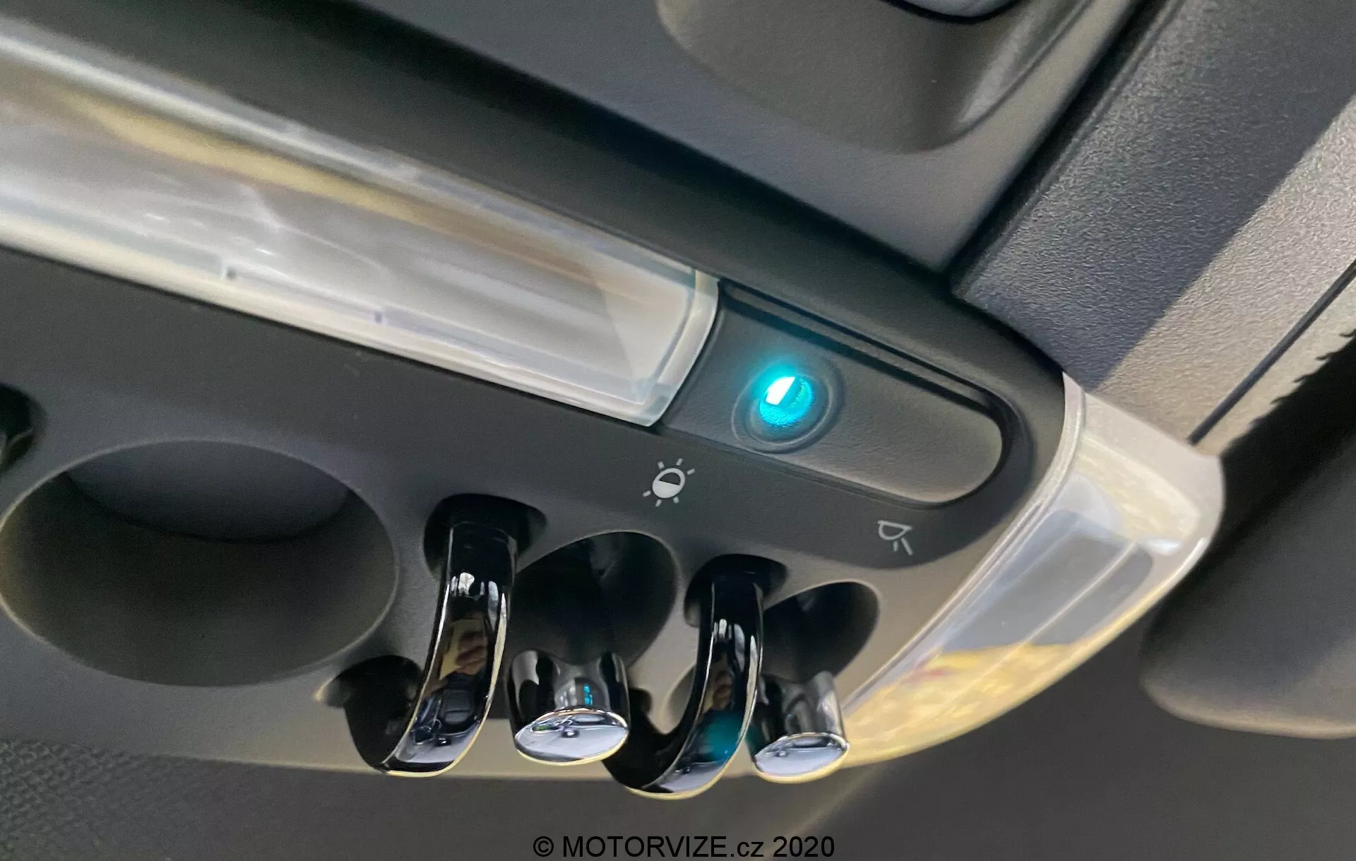 Detailný pohľad na oblasť stropnej konzoly Mini Countryman 2020 John Cooper Works zobrazujúci vnútorné osvetlenie kabíny, tri prepínače s chrómovaným povrchom pre jednotlivé čítacie svetlá, centrálny svetelný prvok kabíny, modré podsvietené tlačidlo pravdepodobne indikujúce Bluetooth alebo bezpečnostnú funkciu, okraj možného úložného priestoru, ikony naznačujúce ďalšie ovládacie prvky alebo senzory a moderný dizajn s lesklými a matnými povrchmi.