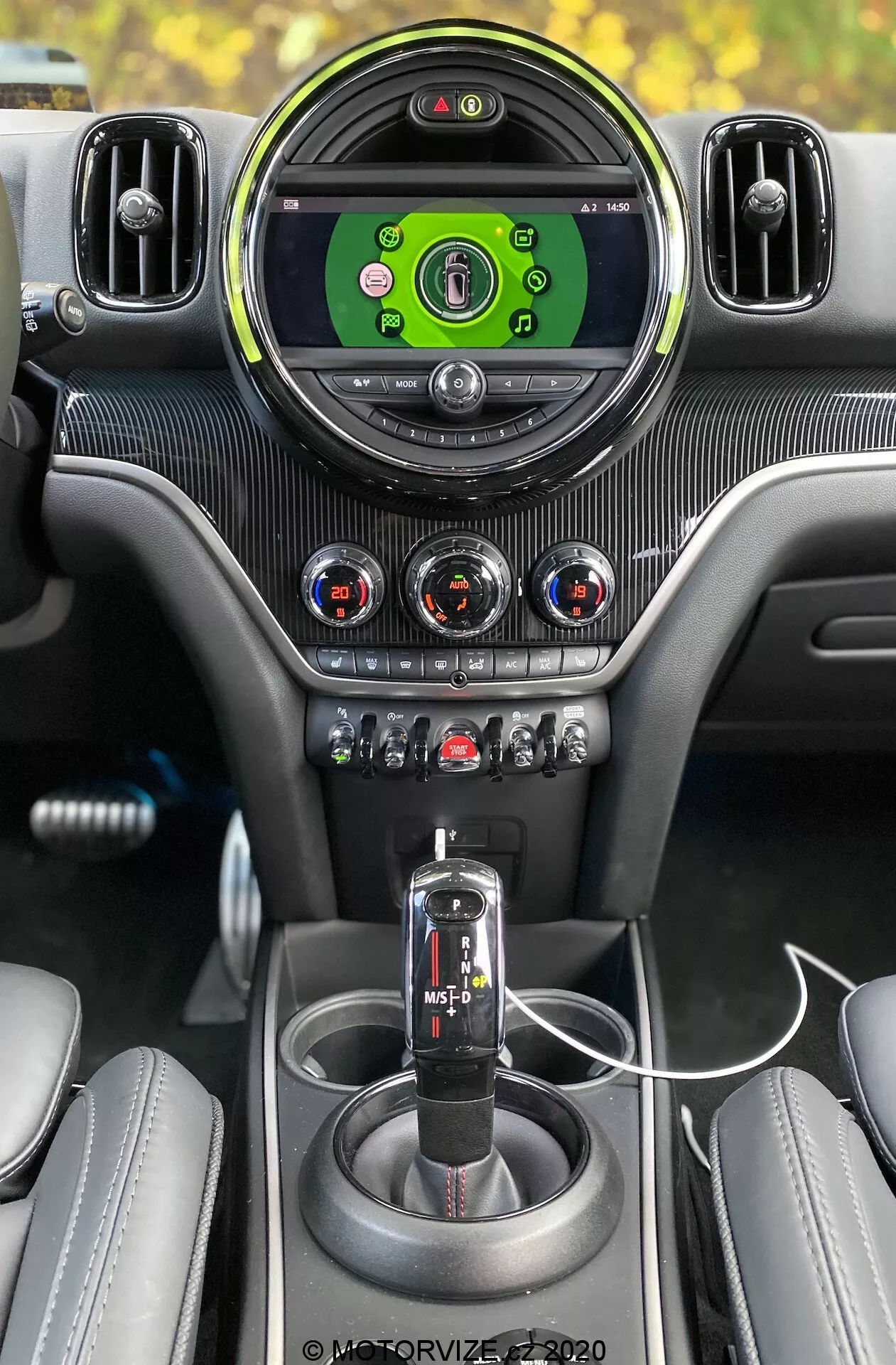 Pohľad_na_interiér_2020_Mini_Countryman_John_Cooper_Works_z_pohľadu_šoféra,_zvýrazňujúci_prémiové_funkcie,_ako_je_veľký_okrúhly_centráný_palubný_displej_s_zeleným_rozhraním_obklopený_striebornými_nastaviteľnými_vetracími_otvormi;_trtjem_dvlôžkové_kujnôpožiisuné_tlačidla_s_červenými_a_modrými_indikátormi_na_karbónovom_vzore_paneli;_rad_tlačidiel_a_spínačov_pre_rôznous_i_funkcie_auta,_vrátane_s_mode_sport/trakcia;_črna_razenná_páka_automotenic_konfi_n_opčnou;_celistvá_středová_konsola_s_karbónovým_vzorom;_dva_drzaky_na_napoje;_pripojený_propojvaci_kábel_USB;a čiastočne_viditeľne_šívané_kožené_sedadlá,_to_všetko_tvorí_prvotriedny_technologicky_zameraný_automobilový_interiér.
