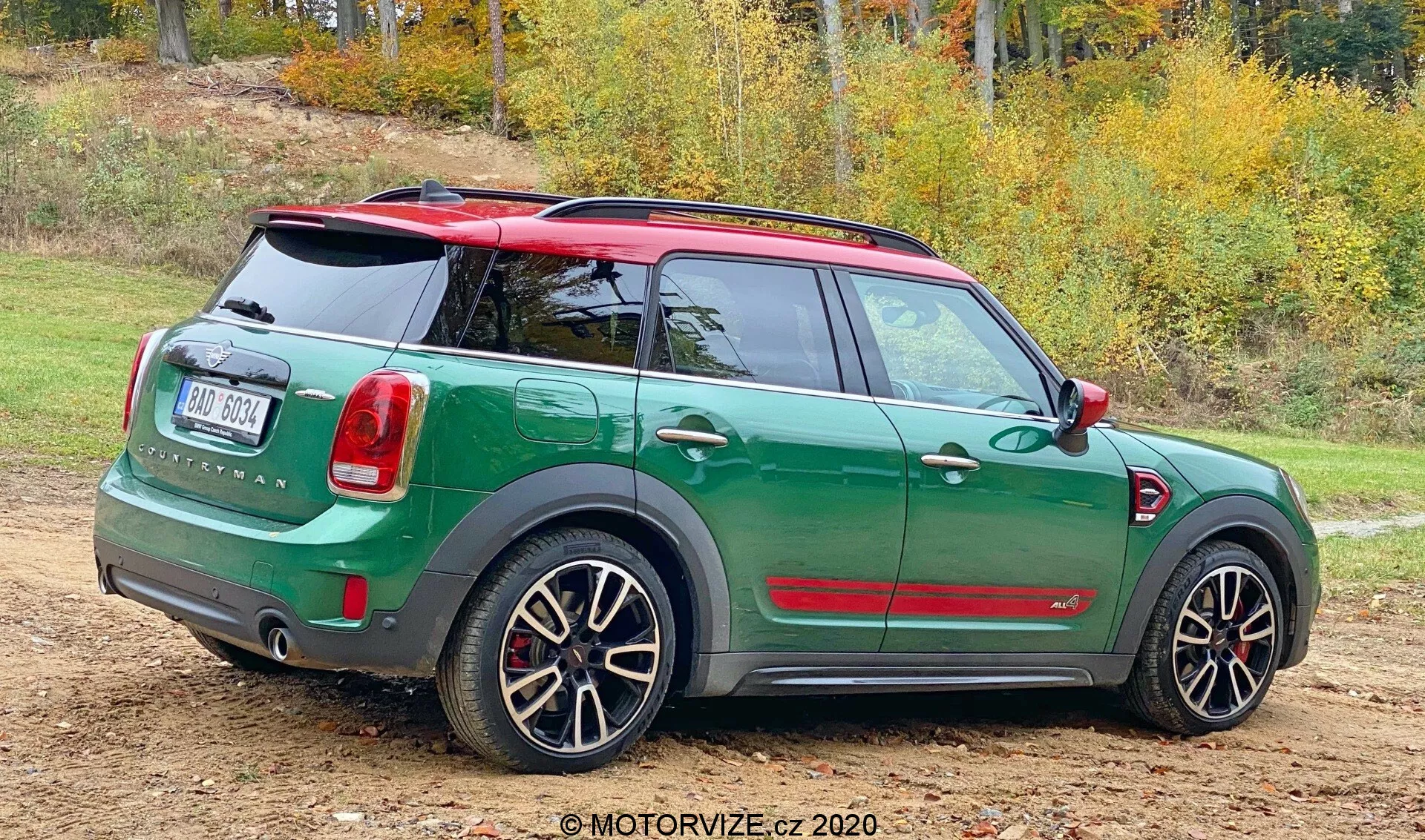 Trojštvrťový zadný pohľad na Mini Countryman John Cooper Works 2020 stojace na nezpevnenom povrchu s miestami trávy. Športové kompaktné auto má dvojfarebnú schému s tmavozelenou karosériou a kontrastnou červenou strechou a zrkadlami. Zadný dizajn ukazuje zadný poklop, zadný stierač, výrazný odznak a vertikálne usporiadané zadné svetlá. Zadný nárazník obsahuje parkovacie senzory. Má tiež športové zliatinové kolesá s viacšpicovým dizajnom, ktoré odhaľujú brzdové strmene, čo naznačuje výkonovú verziu. Prirodzené pozadie s jesennými stromami naznačuje ročné obdobie.
