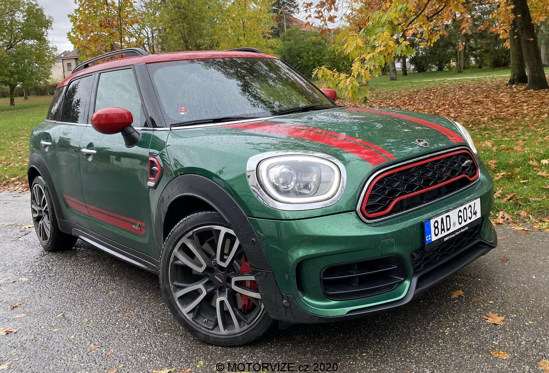 Predný trojštvrťový pohľad na Mini Countryman John Cooper Works z roku 2020, ktorý zobrazuje jeho zelenú kompaktnú SUV karosériu s kontrastnými červenými akcentmi. Zachytený z nízkeho uhlu, obraz zvýrazňuje športovú prednú mriežku s chrómovým rámom a včelím plástovým dizajnom, výrazné kruhové svetlomety s LED dennými svetlami, viac lúčové zliatinové disky kolies so šedým povrchom a červené kryty zrkadiel. Fotografia je nastavená na pozadí stromov a slabej štruktúry, čo naznačuje umiestnenie v parku alebo rezidenčnej oblasti.