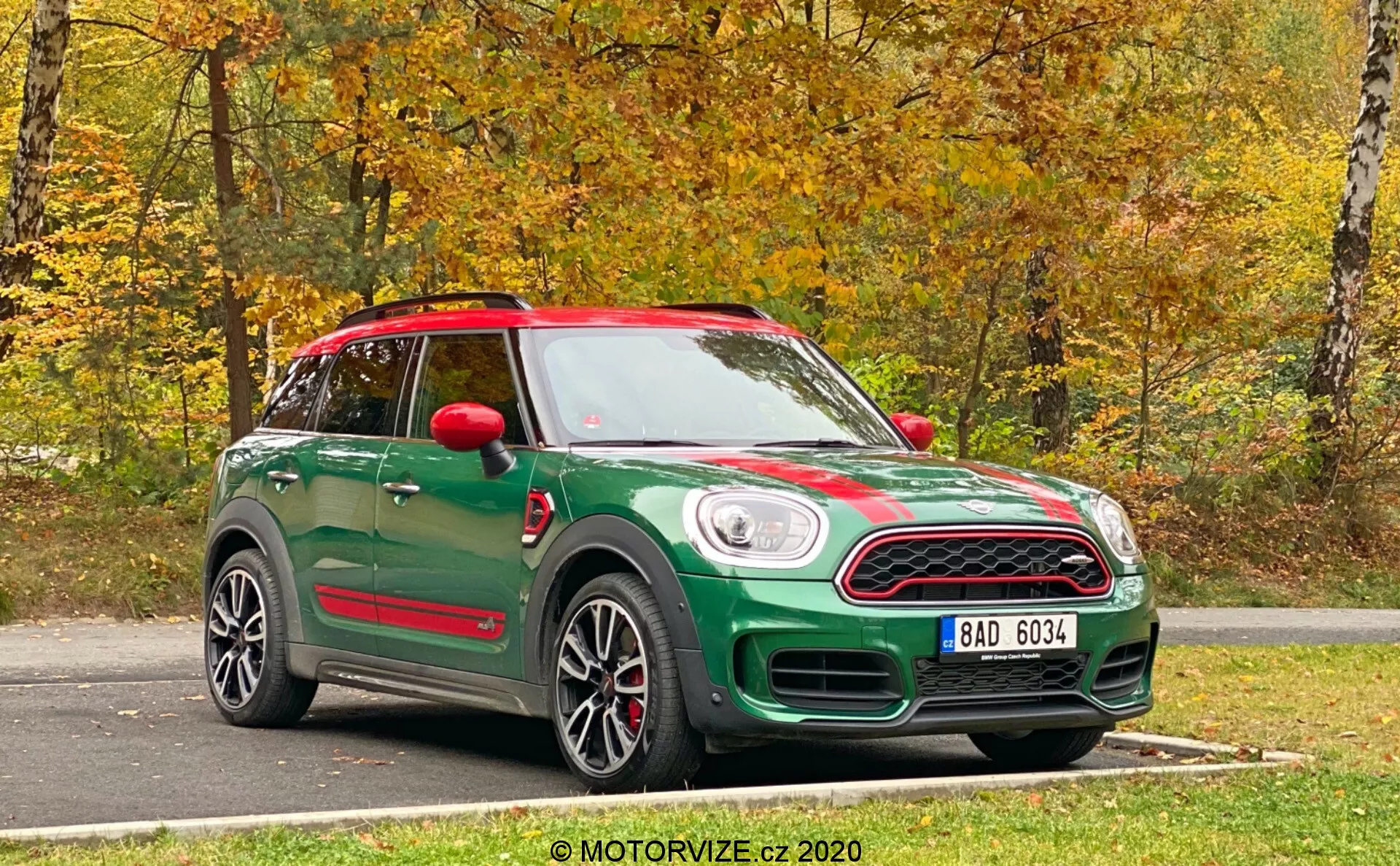 Zelený Mini Countryman John Cooper Works 2020 zachytený z predného trojštvrťového pohľadu. Fotografia ukazuje prednú časť aj bočný profil auta, zdôrazňujúc jeho dizajnové prvky. Predná časť má výraznú mriežku s evidenčným číslom pod ňou a klasické okrúhle svetlomety. Na kapote bežia športové pruhy a bočný profil ukazuje zliatinové kolesá, bočné zrkadlá a líniu strechy. Červené bočné zrkadlá a zladená strecha vytvárajú dvojfarebnú farebnú schému. Pod dverami farebne zladené bočné prahy naznačujú vyšší stupň výbavy alebo športový balíček. Vozidlo má panoramatické strešné okno a je zaparkované na asfalte s prírodným jesenným pozadím. Ostrá, jasná fotka bola urobená za prirodzeného denného svetla.