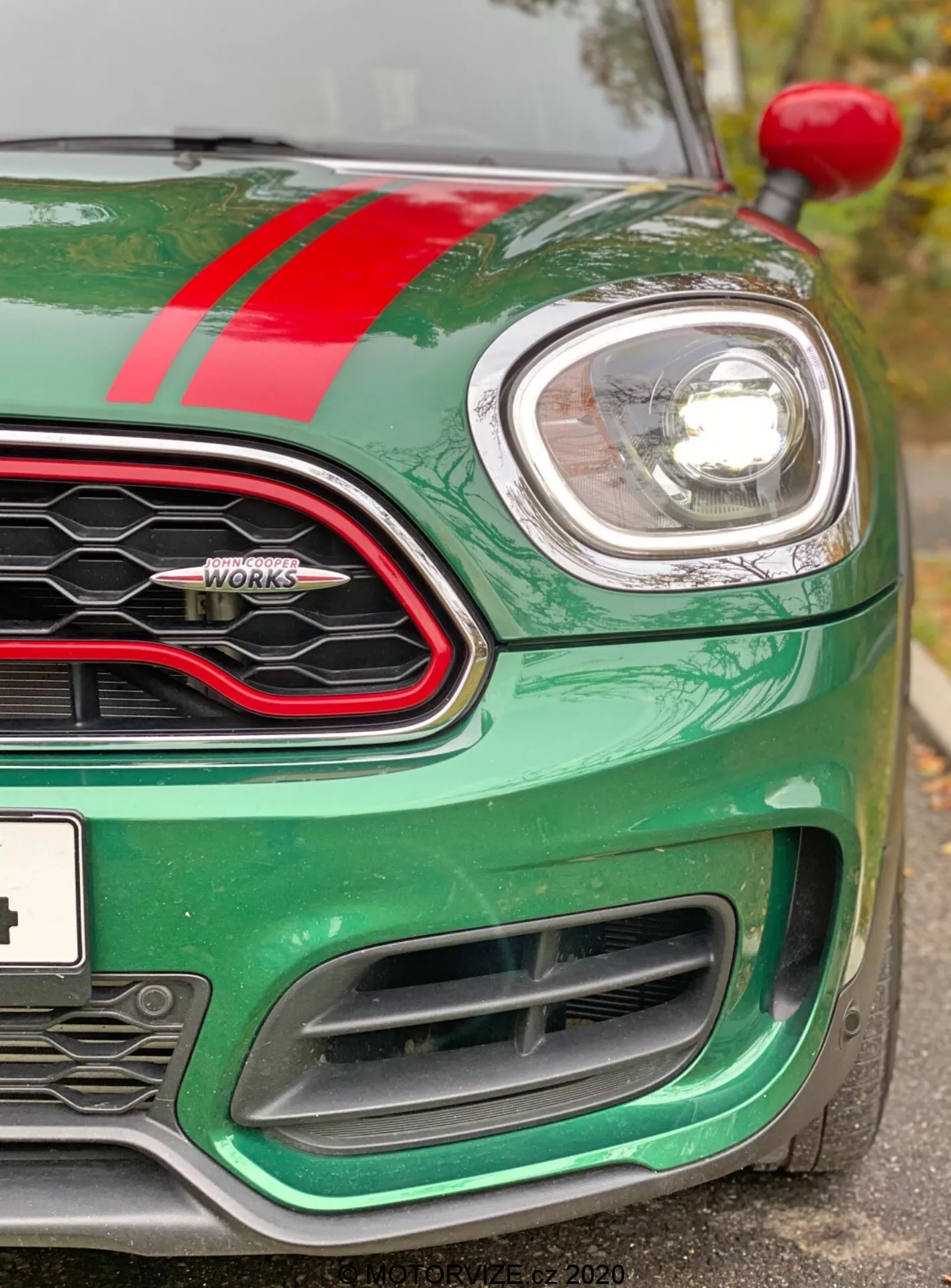 Detailný pohľad na prednú ľavú štvrtinu vozidla Mini Countryman John Cooper Works z roku 2020. Snímka zhotovená mierne pod úrovňou svetlometov zvýrazňuje kovovo zelenú kapotu s červeným závodným pruhom, kruhový ľavý svetlomet uzavretý v chróme a časť mriežky vo tvare včelieho plástu s lesklým čiernym rámom. Viditeľný je znak 'John Cooper Works', ľavý indikátor na oblúku kolesa a veľké nasávacie otvory predného nárazníka navrhnuté na chladenie motora a aerodynamický výkon. Auto vyzerá čisto a dobre udržiavané, zdôrazňujúc modernú techniku svetlometov s projektorom alebo LED a športový, výkonnostne orientovaný dizajn.