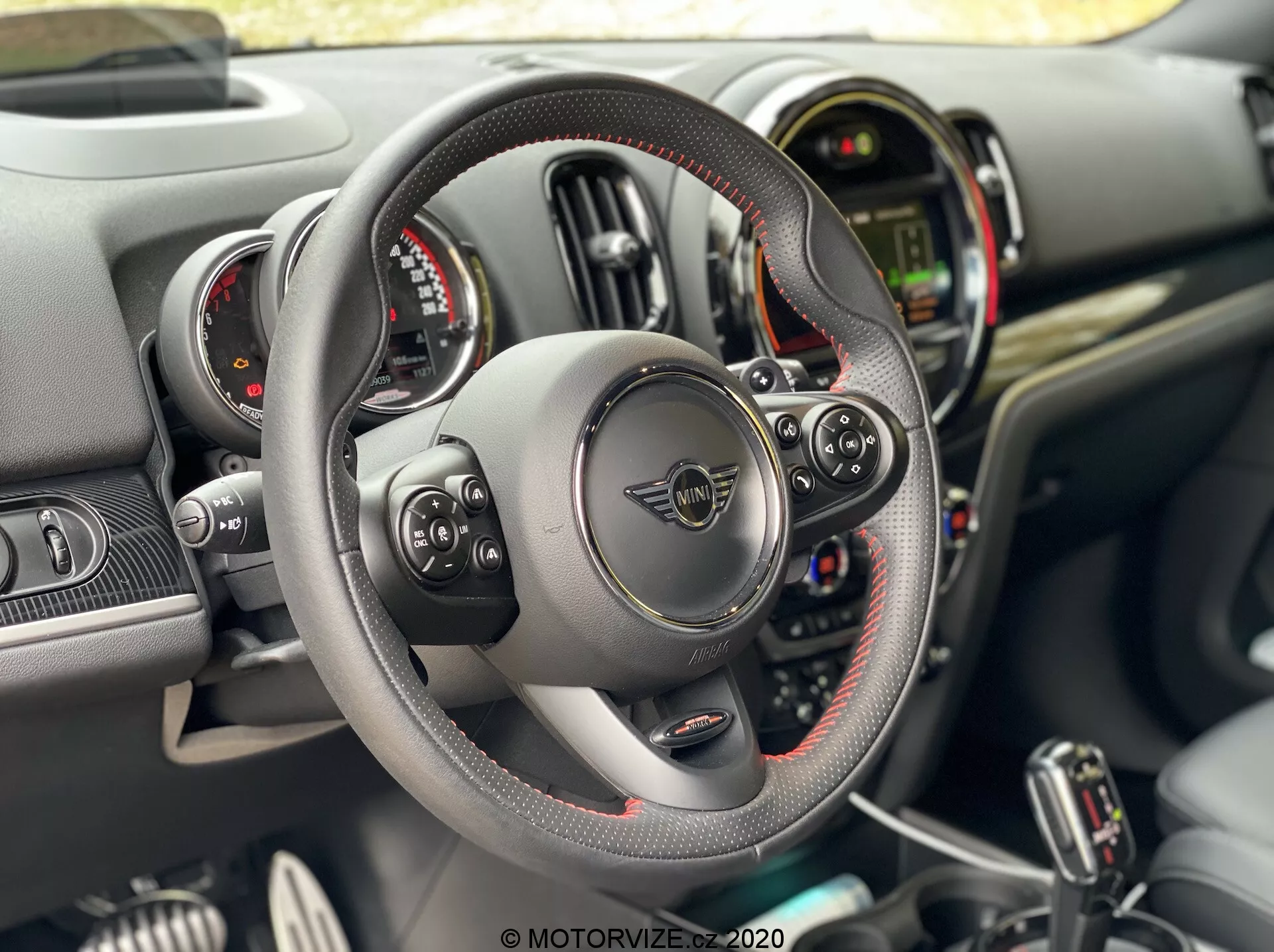Interiér vozidla Mini Countryman John Cooper Works 2020 z pohľadu vodiča s koženým volantom s plochým dnom, viditeľným šitím a multifunkčnými ovládačmi, pákami radenia za volantom, dvojitými kruhovými ciferníkmi prístrojov obklopujúcimi centrálny digitálny displej a palubnou doskou s rôznymi tlačidlami a prepínačmi na stredovej konzole.